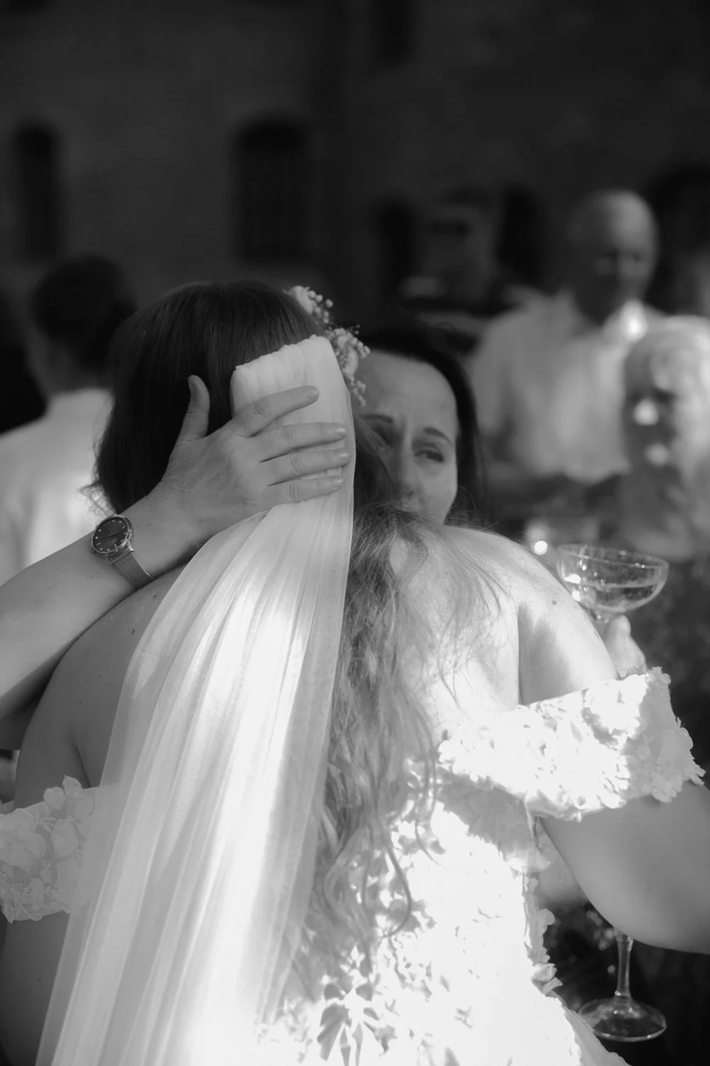 Two women hugging at a wedding celebration, one wearing a wedding dress and veil, the other in a dress, with several guests visible in the background.