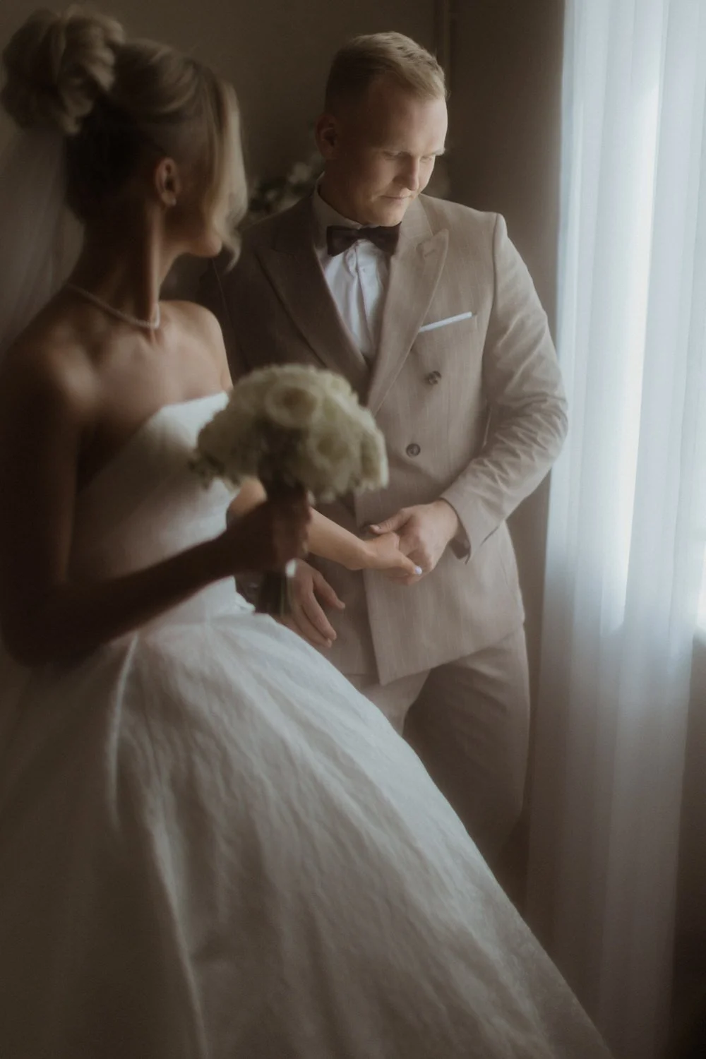 A bride and groom standing near a window, holding hands, with the bride holding a bouquet of white roses.