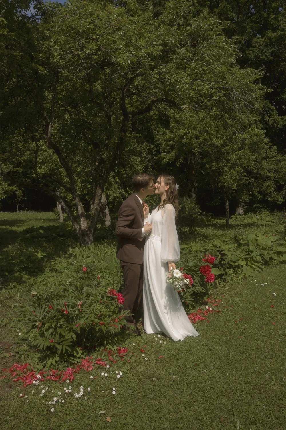 A couple in wedding attire standing outdoors in a green garden, sharing a kiss, with the woman holding a bouquet of flowers.