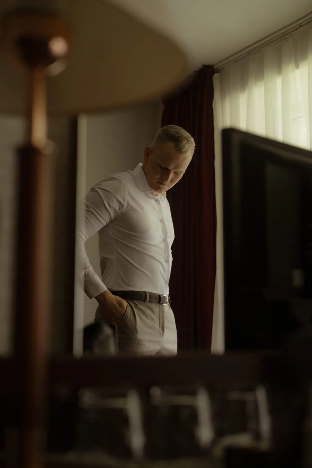 A man in a white shirt and beige pants stands with his hand in his pocket, looking down thoughtfully in a dimly lit room with curtains and a lamp.