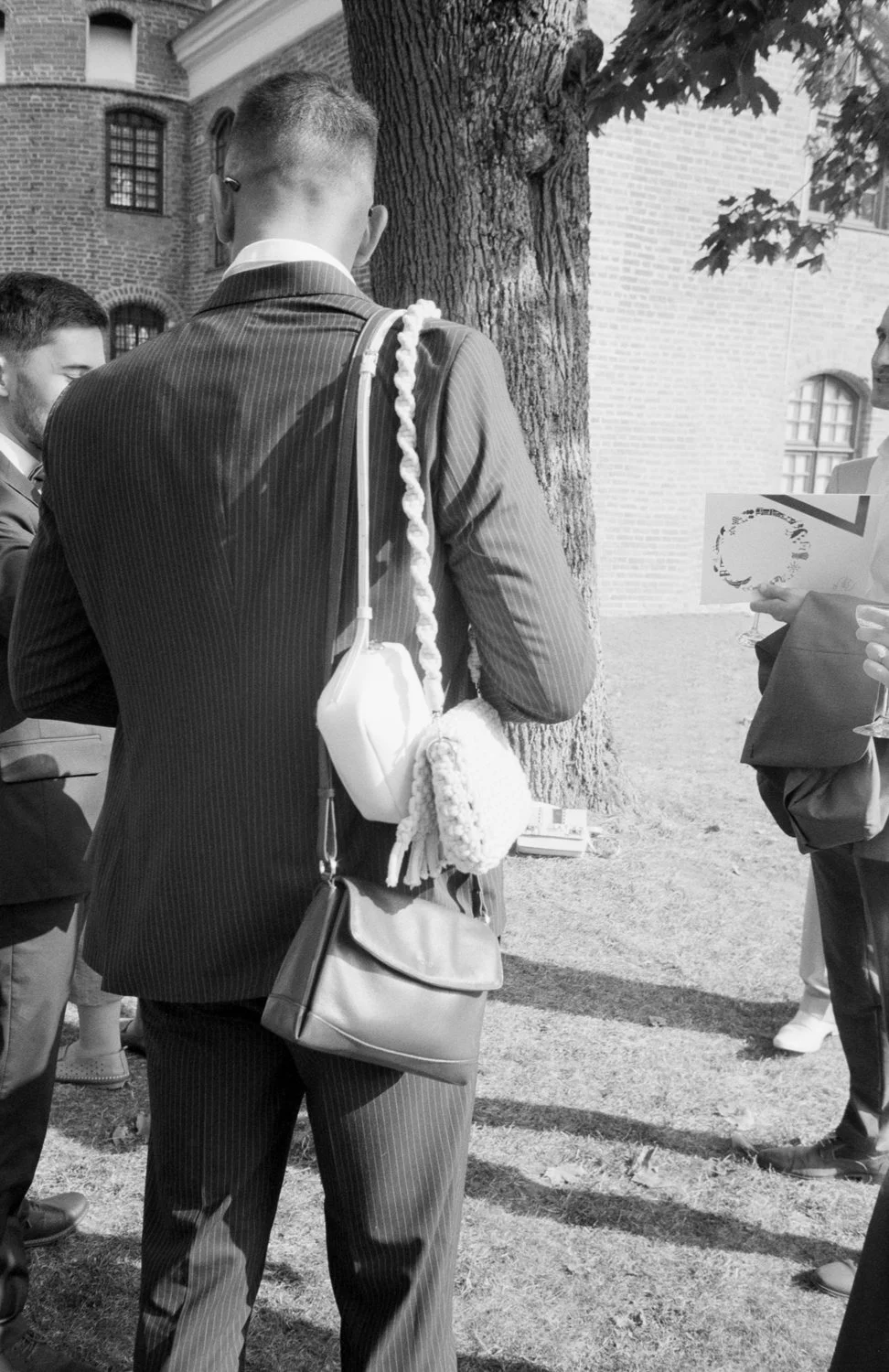 Men in suits gathered outdoors near a brick building and a large tree, engaging in conversation