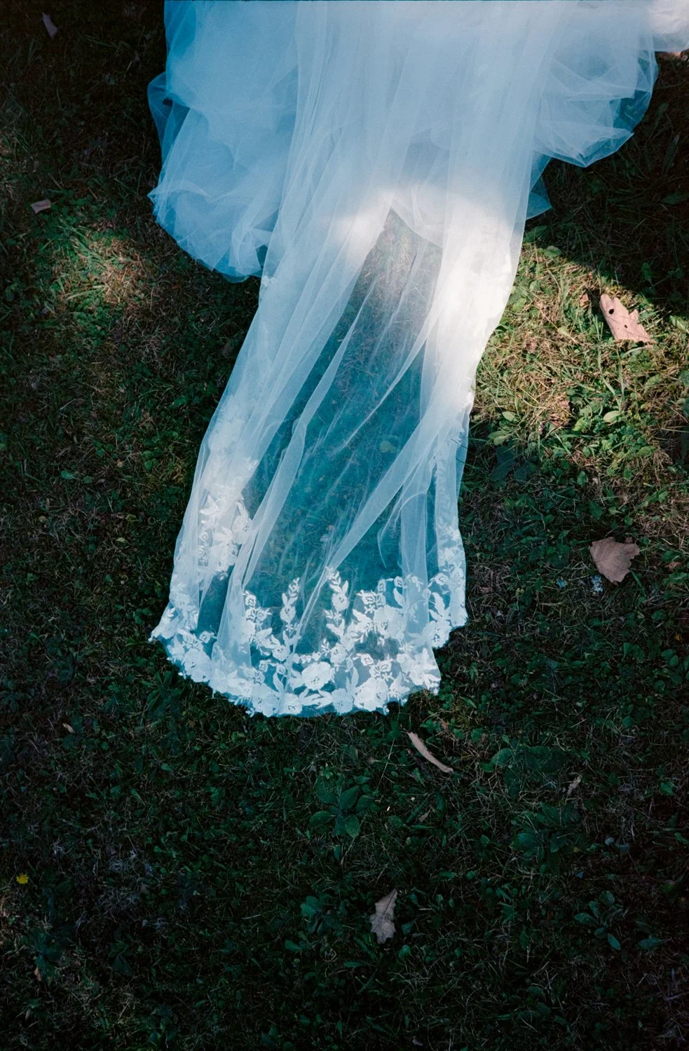 A long white wedding veil with embroidered floral edges lying on grass with leaves around.