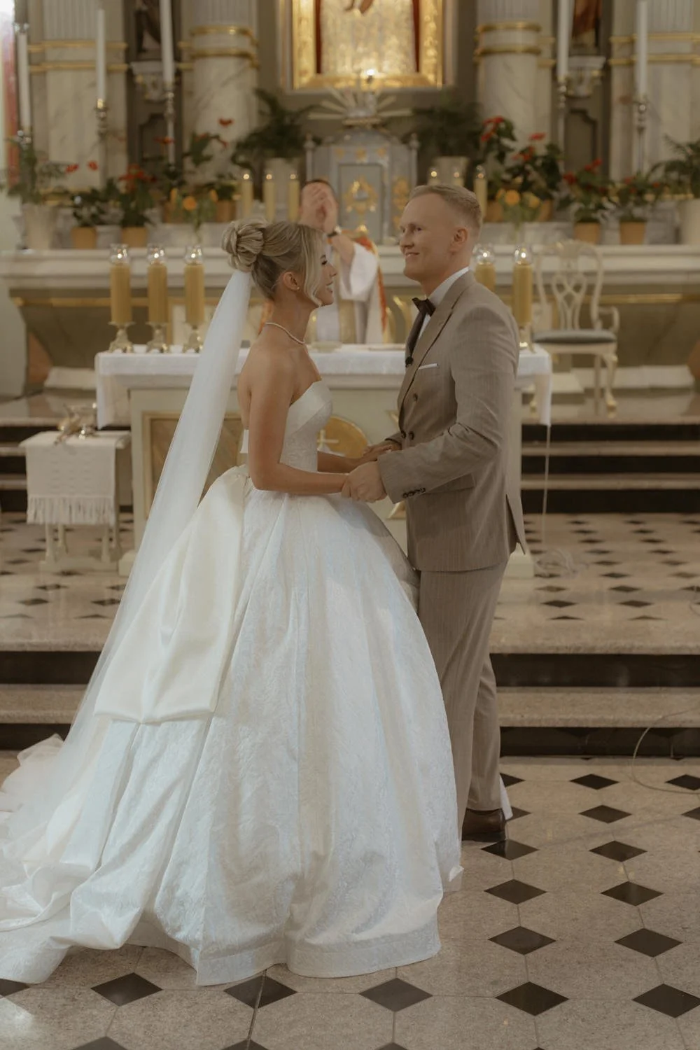 A bride and groom holding hands during their wedding ceremony inside a church, with an officiant in the background.