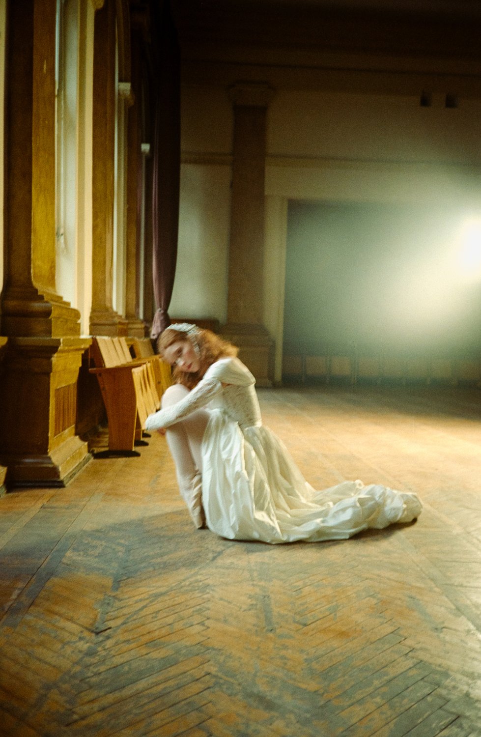 A woman in a white wedding dress sitting on the wooden floor of an empty, dimly lit room, with her head resting on her knees, and chairs along the wall behind her.