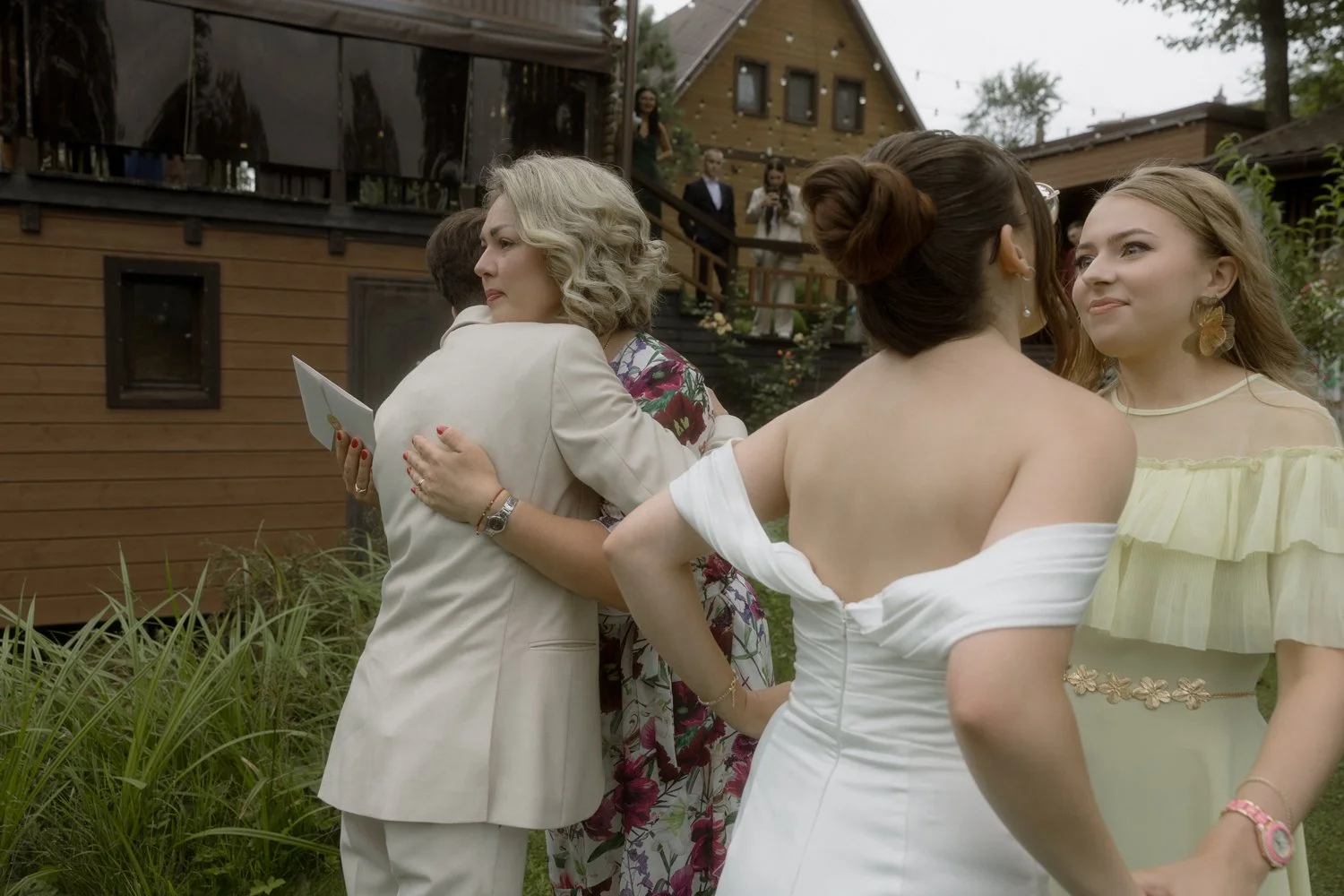 A group of women at an outdoor event, hugging and talking in a garden area with wooden buildings in the background.