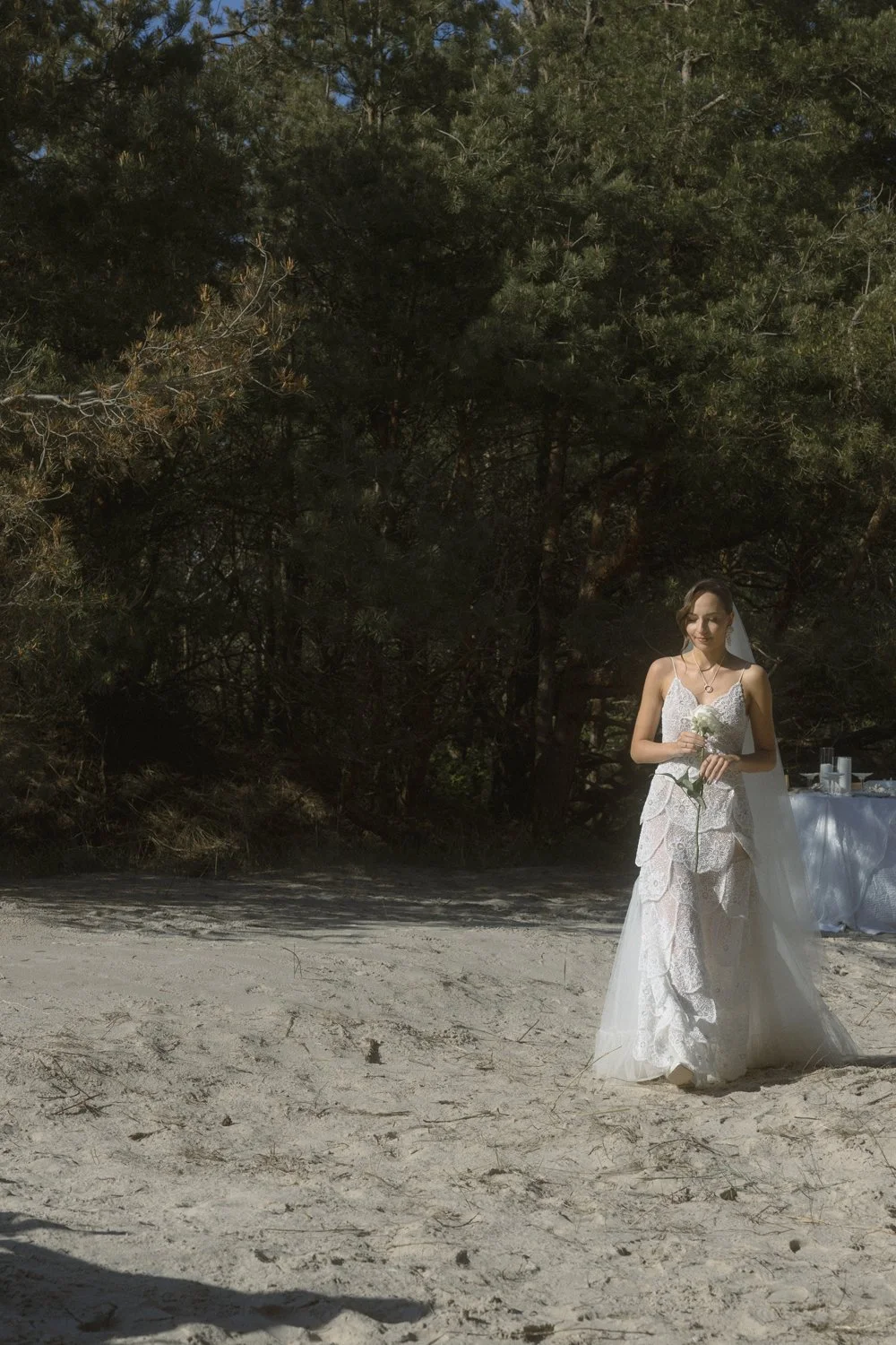 A bride in a white lace wedding dress holding a white flower, standing on a sandy beach with trees in the background.