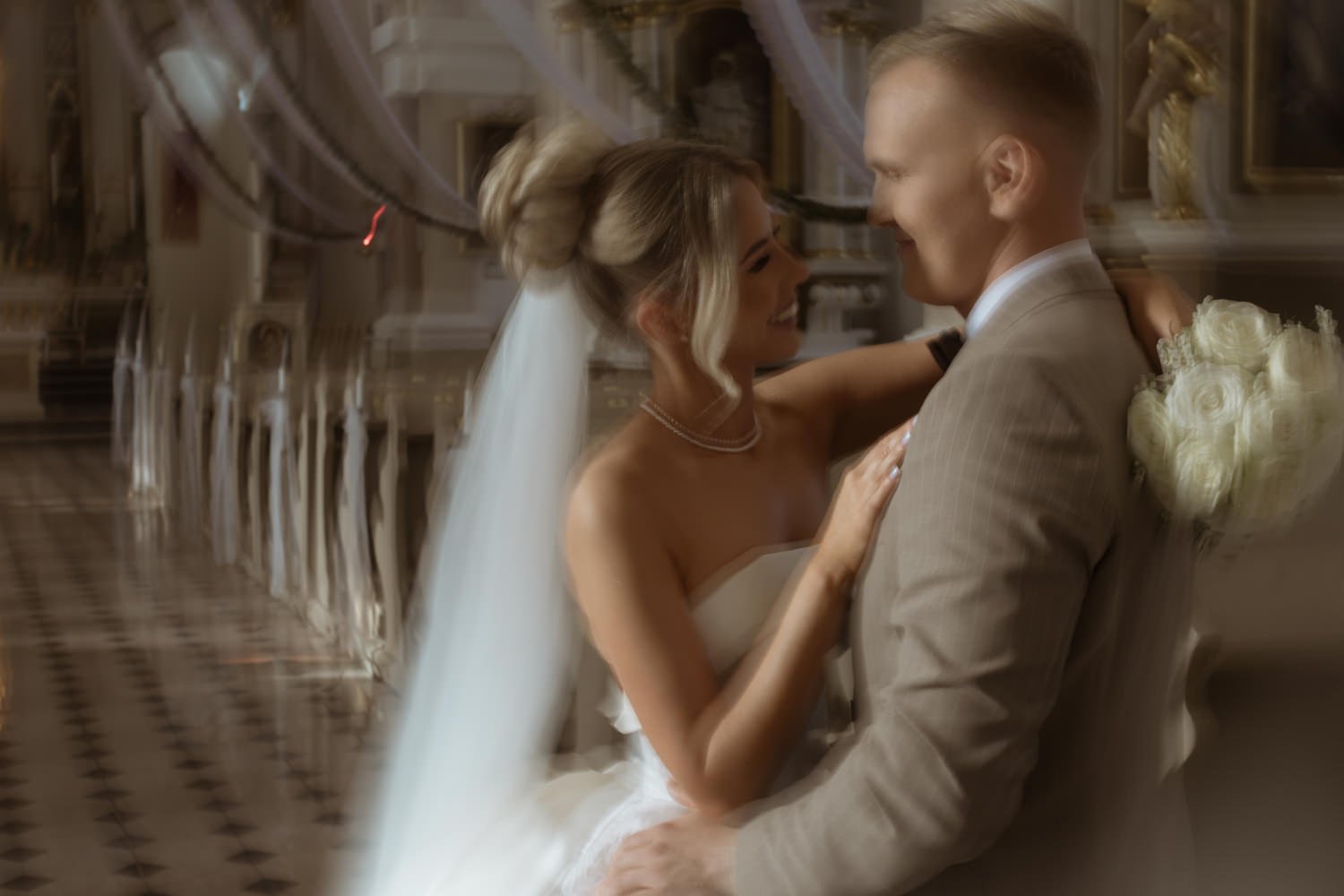 A bride and groom smiling and standing close together indoors, with the bride wearing a white wedding dress and veil, and the groom in a light-colored suit. The bride has her hands on the groom's chest, and the groom holds a bouquet of white roses be