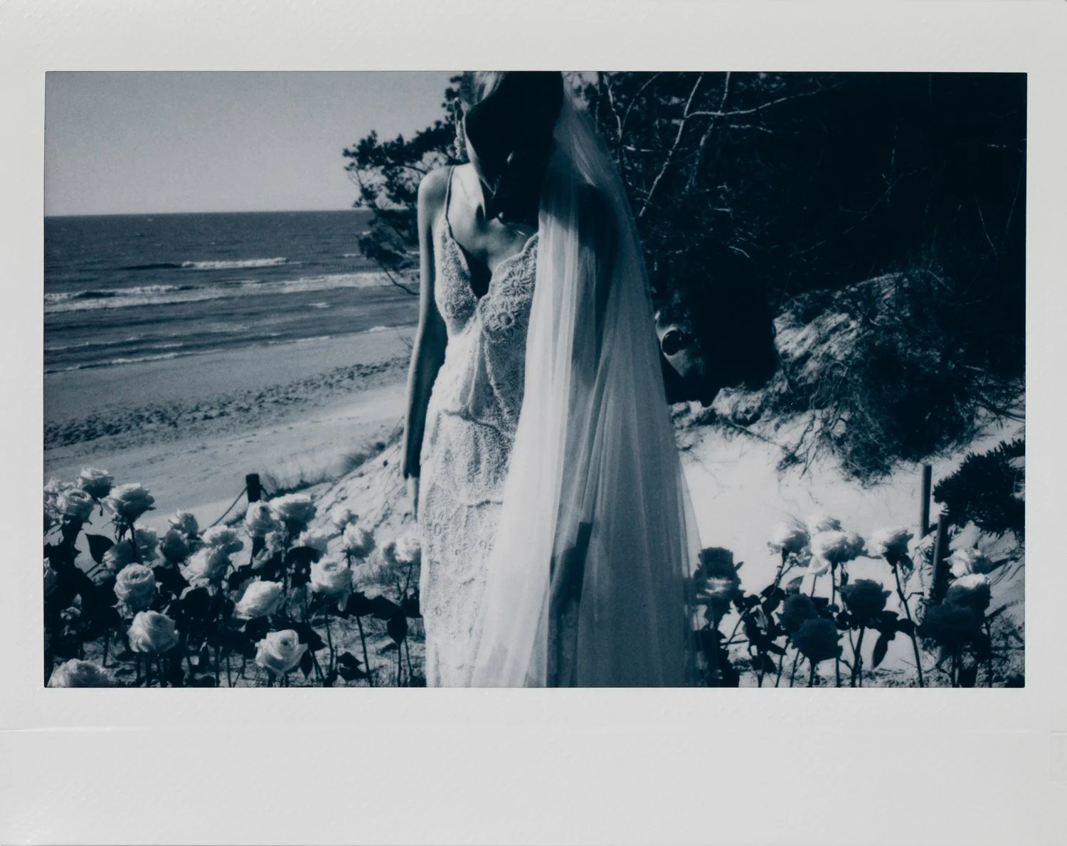 Black and white photo of a bride in a lace dress and veil standing on a beach with flowers in the foreground and ocean waves in the background.