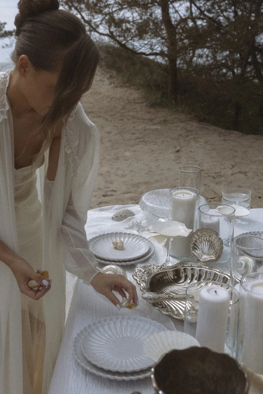 A woman arranging a table with white and silver dishes, candles, and seashells outdoors on a sandy beach with trees in the background.