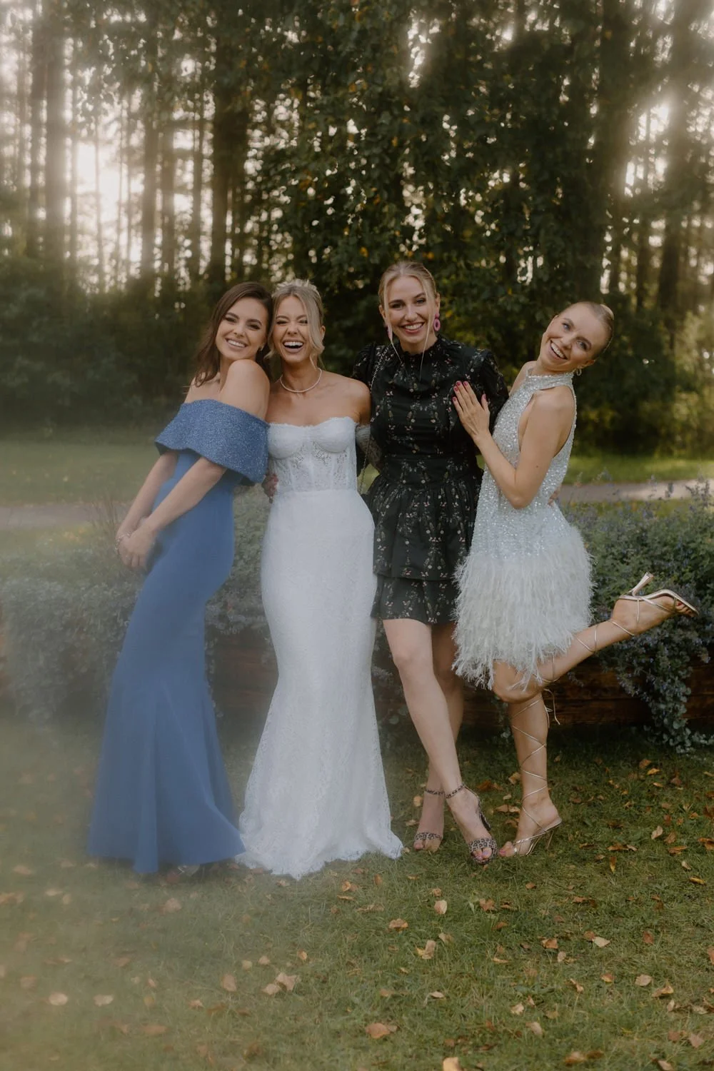 Four women wearing formal dresses standing together outdoors in a park surrounded by trees and bushes, smiling and posing for a photo.