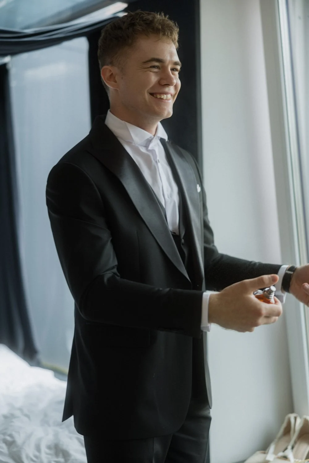 A young man in a black tuxedo with a white shirt smiling and holding a perfume bottle while standing by a window.