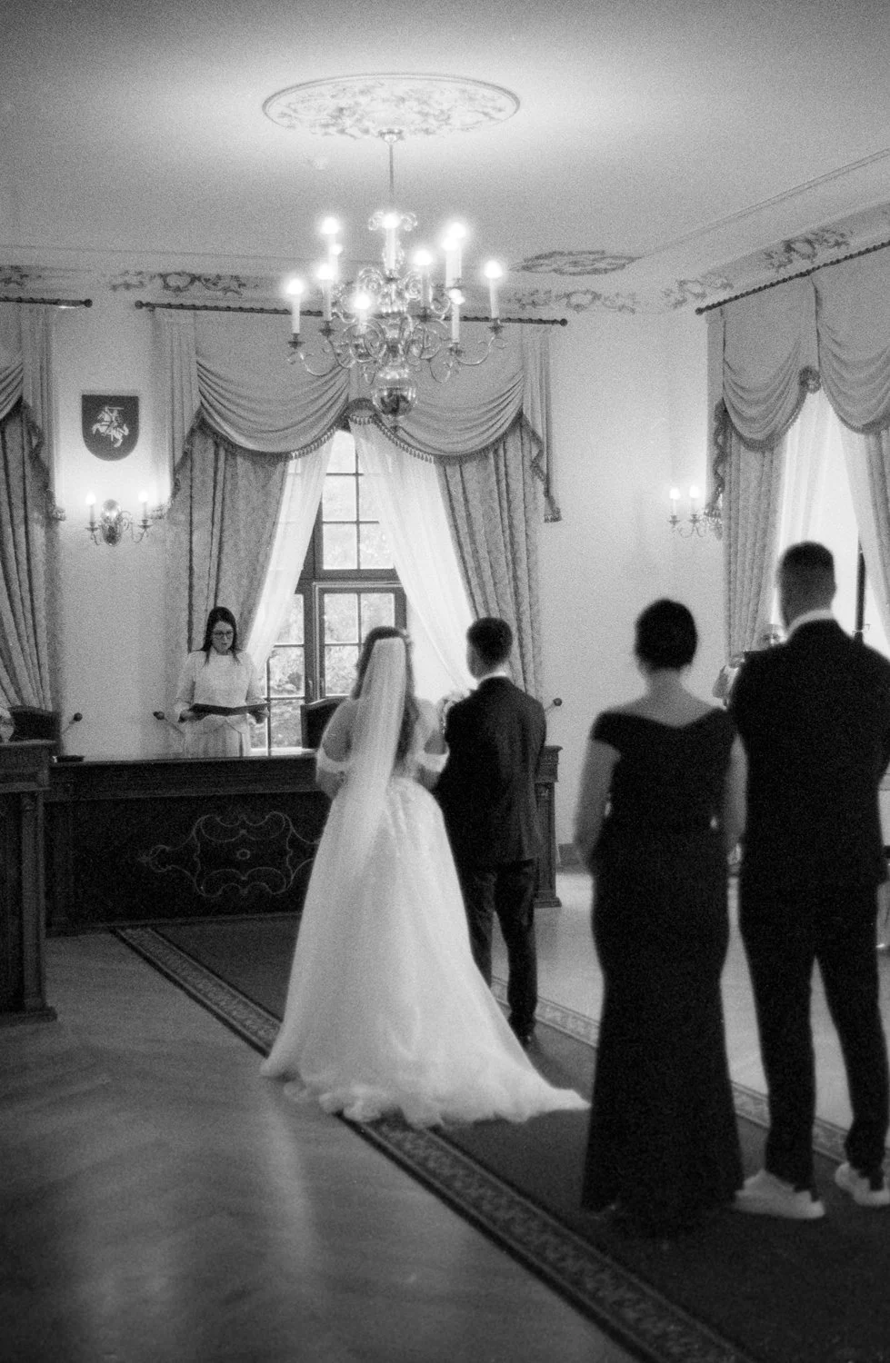 A wedding ceremony taking place in an elegant, historic room with ornate curtains, chandeliers, and a portrait on the wall. The bride and groom stand before an officiant, with two witnesses observing.
