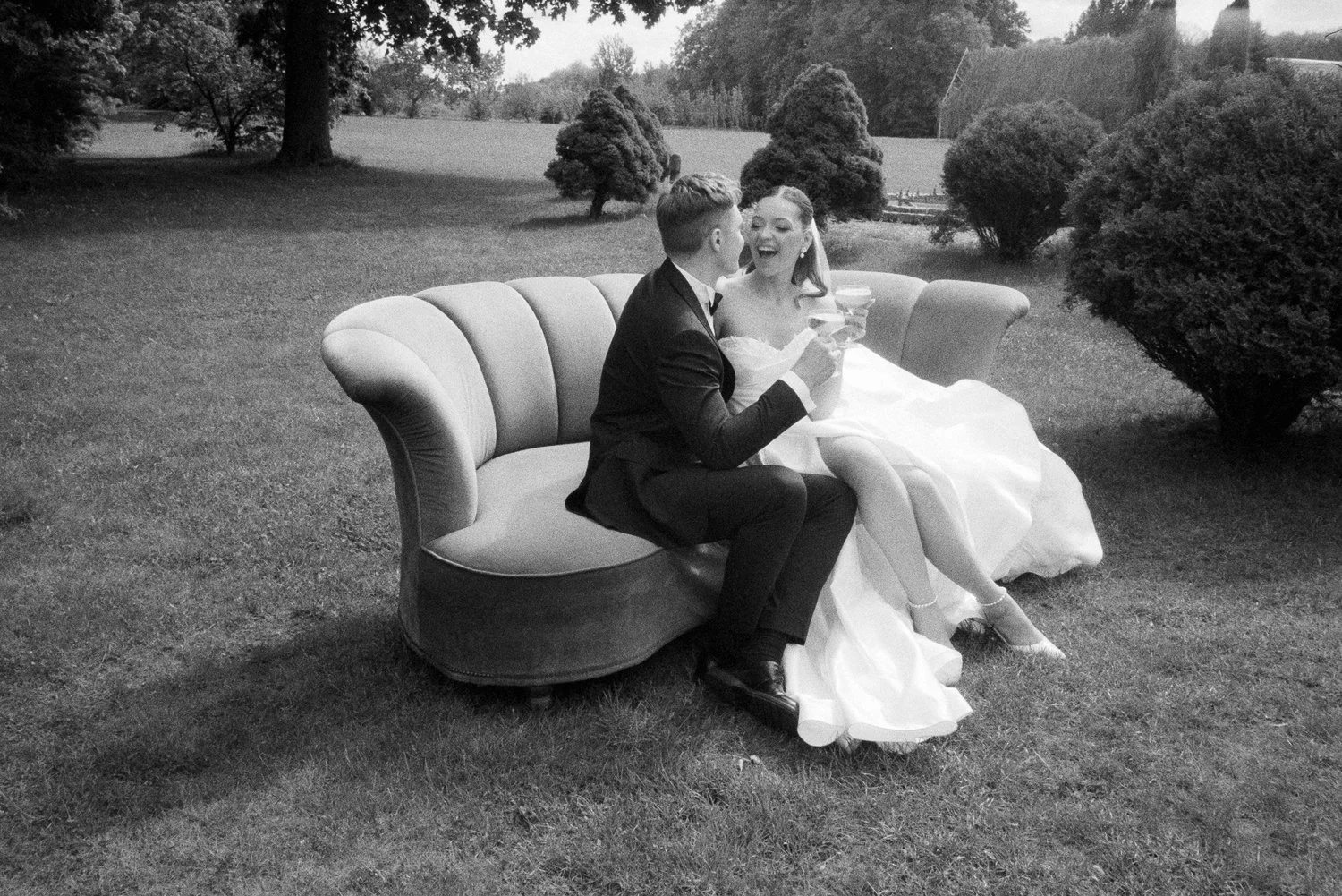 A black-and-white photo of a bride and groom sitting on a vintage sofa outdoors, celebrating with drinks, smiling and engaging with each other, surrounded by trees and bushes.