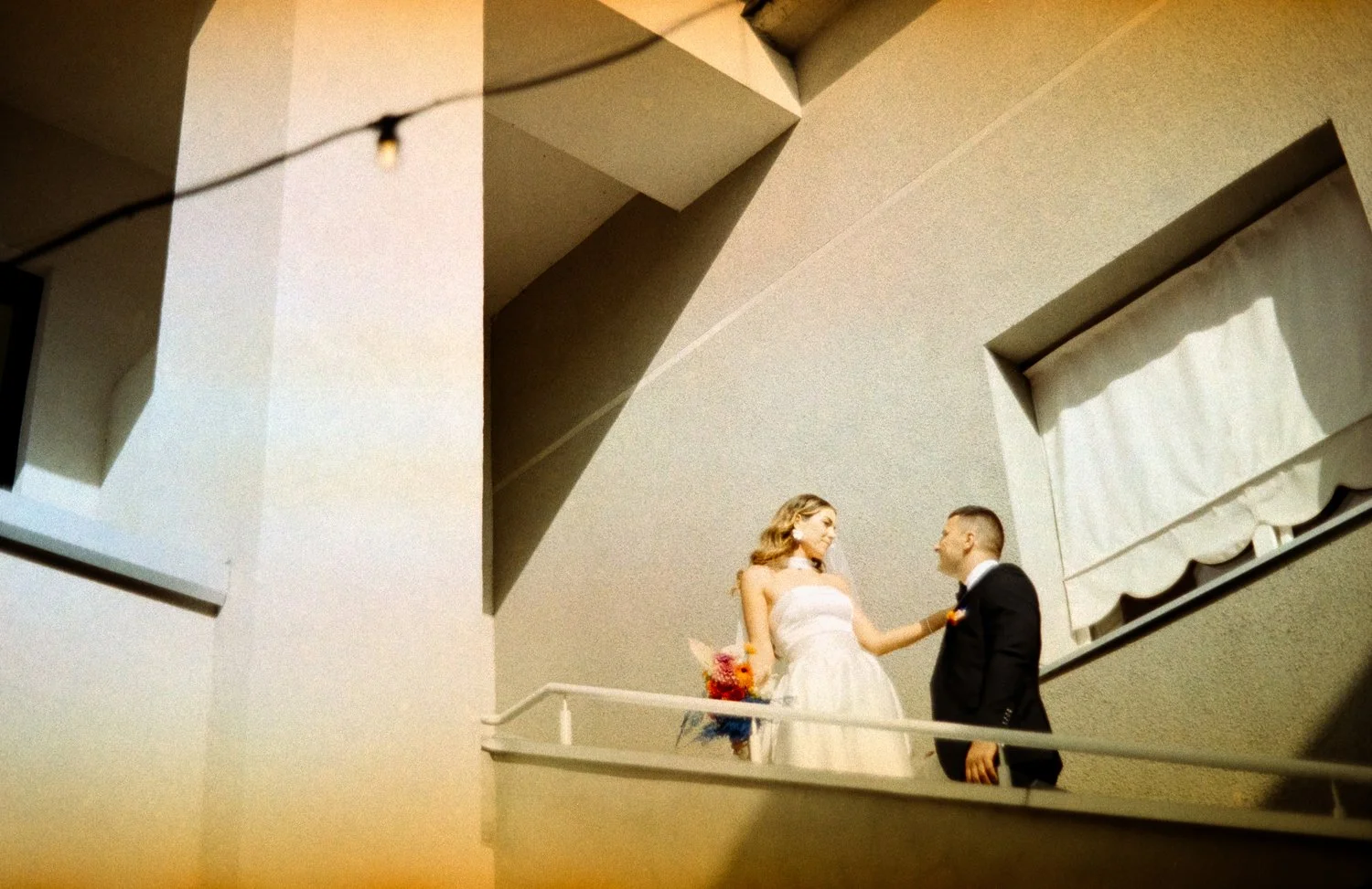 A bride and groom on a balcony, with the bride holding a bouquet of flowers and the groom wearing a black tuxedo.