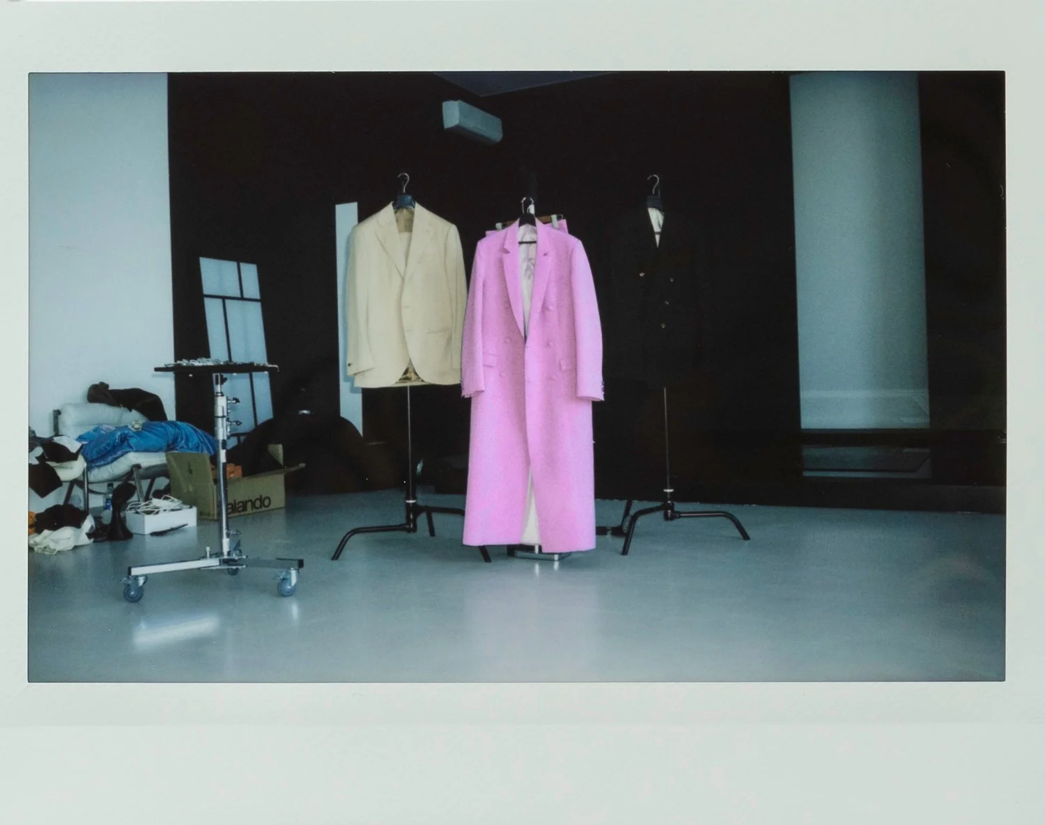 Three blazers hanging on racks, two of which are beige and black, and one pink. The room has a black wall and a window with a black frame. Items like clothing and boxes are on the floor to the left.