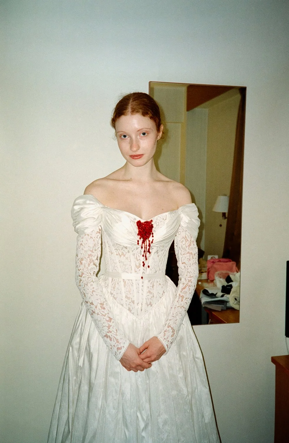 A woman with red hair wearing a white lace off-shoulder dress with red bloodstain details on the chest, standing in a room with a mirror and some furniture in the background.