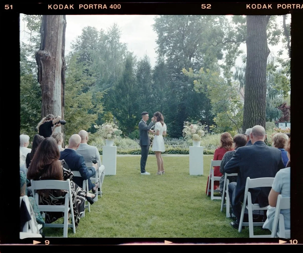 A couple getting married outdoors in a garden, with guests seated in white chairs watching. The officiant or officiant's assistant is taking a photo on the left. The scene is framed by large trees and greenery, creating a natural backdrop.