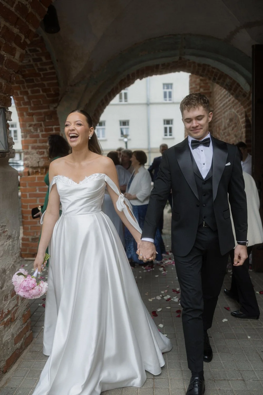 A bride and groom walking hand in hand, smiling and laughing, after their wedding. The bride is wearing a white gown and holding a bouquet of pink flowers; the groom is in a black tuxedo with a bow tie. They are under a brick archway, surrounded by w