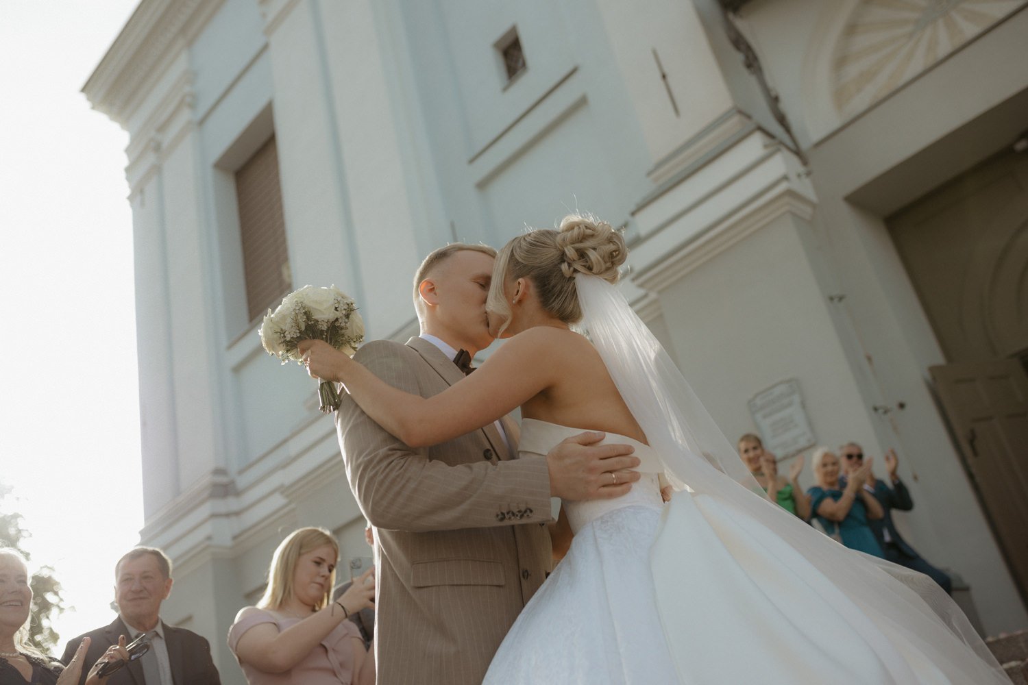 A newlywed couple sharing a kiss outside a church, surrounded by wedding guests taking photos and celebrating.