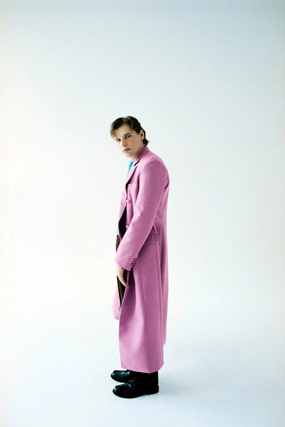 Young man standing in a minimal white studio wearing a pink long coat and black shoes, looking serious.