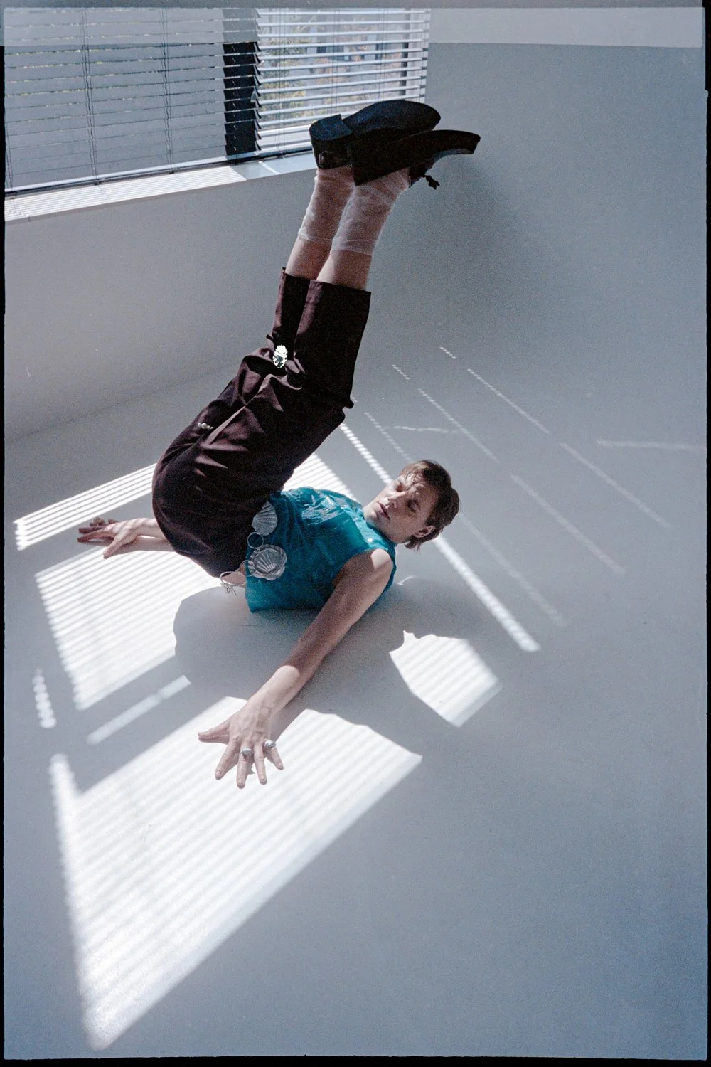 A person wearing a blue sleeveless top and black pants lying on the floor in front of a window with blinds, casting striped shadows, with their right arm extended and legs raised upward, wearing black shoes.