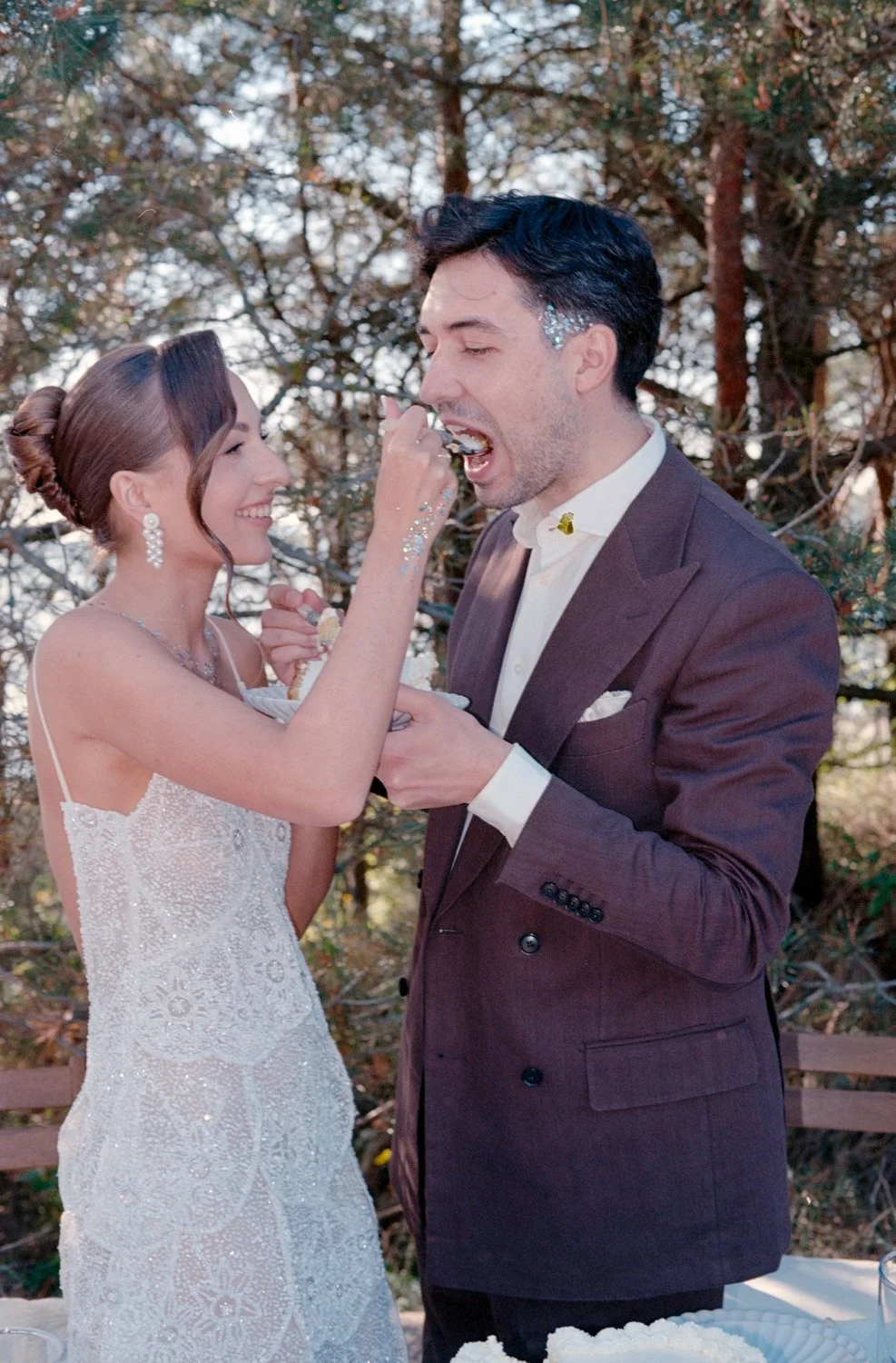 A bride and groom celebrate their wedding by feeding each other cake outdoors, with trees in the background.
