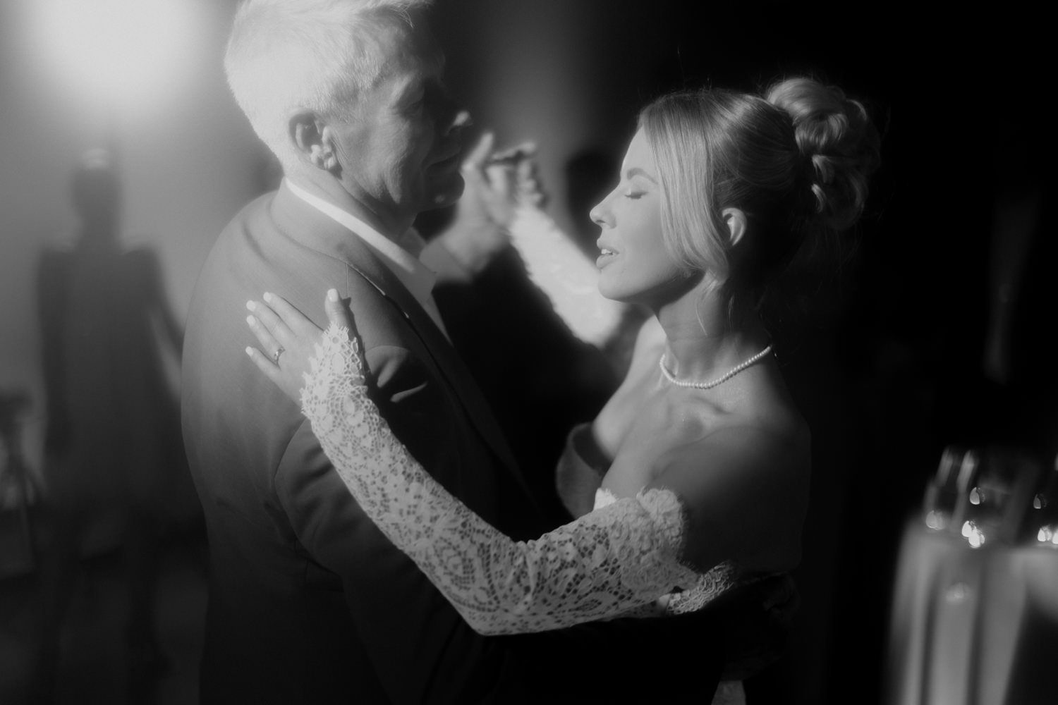 A black and white photo of a bride and an older man dancing closely. The bride is wearing a pearl necklace and a wedding gown with lace sleeves, and the older man is dressed in a suit. They are smiling and appear to be enjoying a special moment.