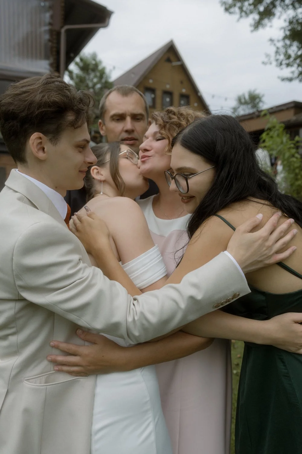 Group of five people hugging, one woman kissing another woman on the cheek, and one man smiling, outdoors with houses and trees in the background.