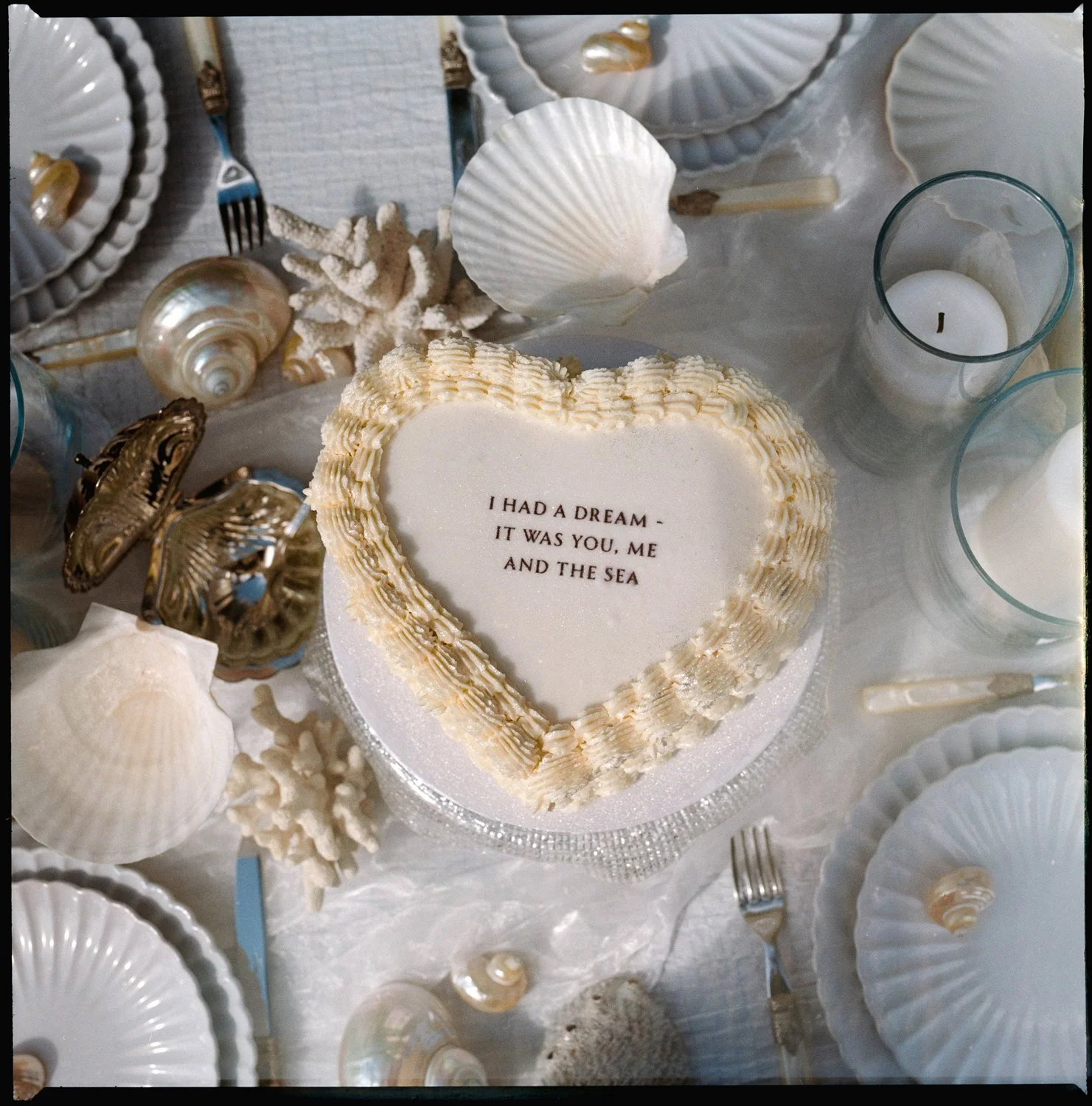 A heart-shaped weddings cake with the message "I HAD A DREAM - IT WAS YOU, ME AND THE SEA" surrounded by seashells, starfish, and candles on a white tablecloth.