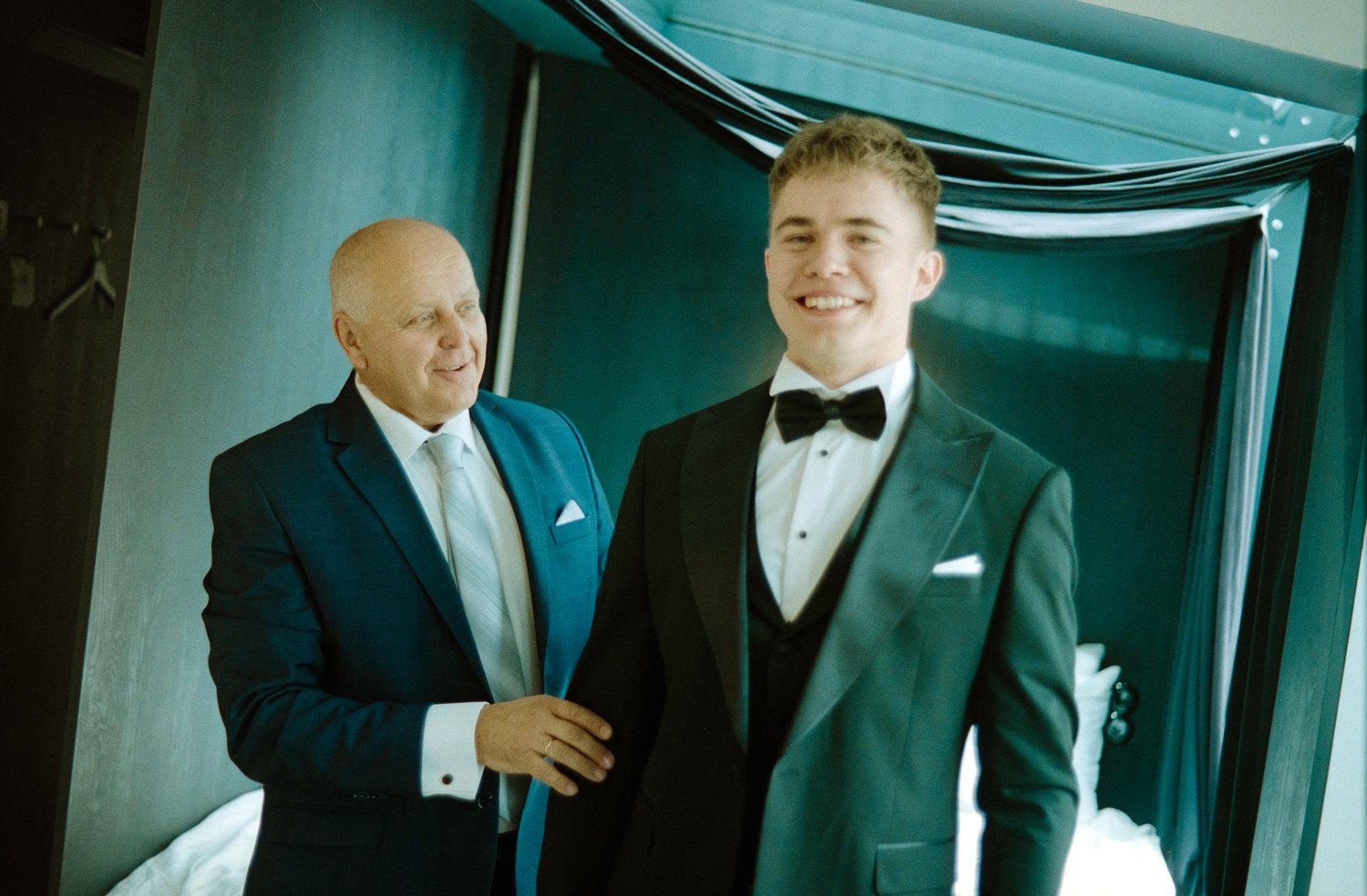 A young man in a tuxedo smiling as an older man, dressed in a suit, adjusts his cufflinks in a hotel room.