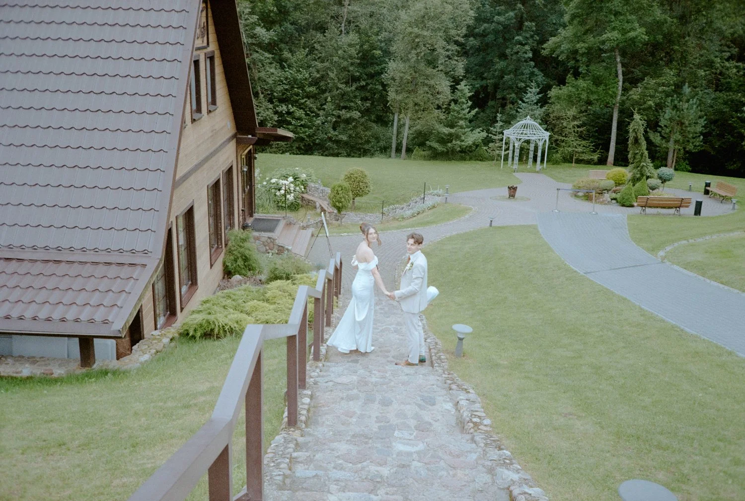 A bride and groom holding hands on a stone pathway outdoors, with a house on the left and a landscaped garden with benches and a gazebo in the background.