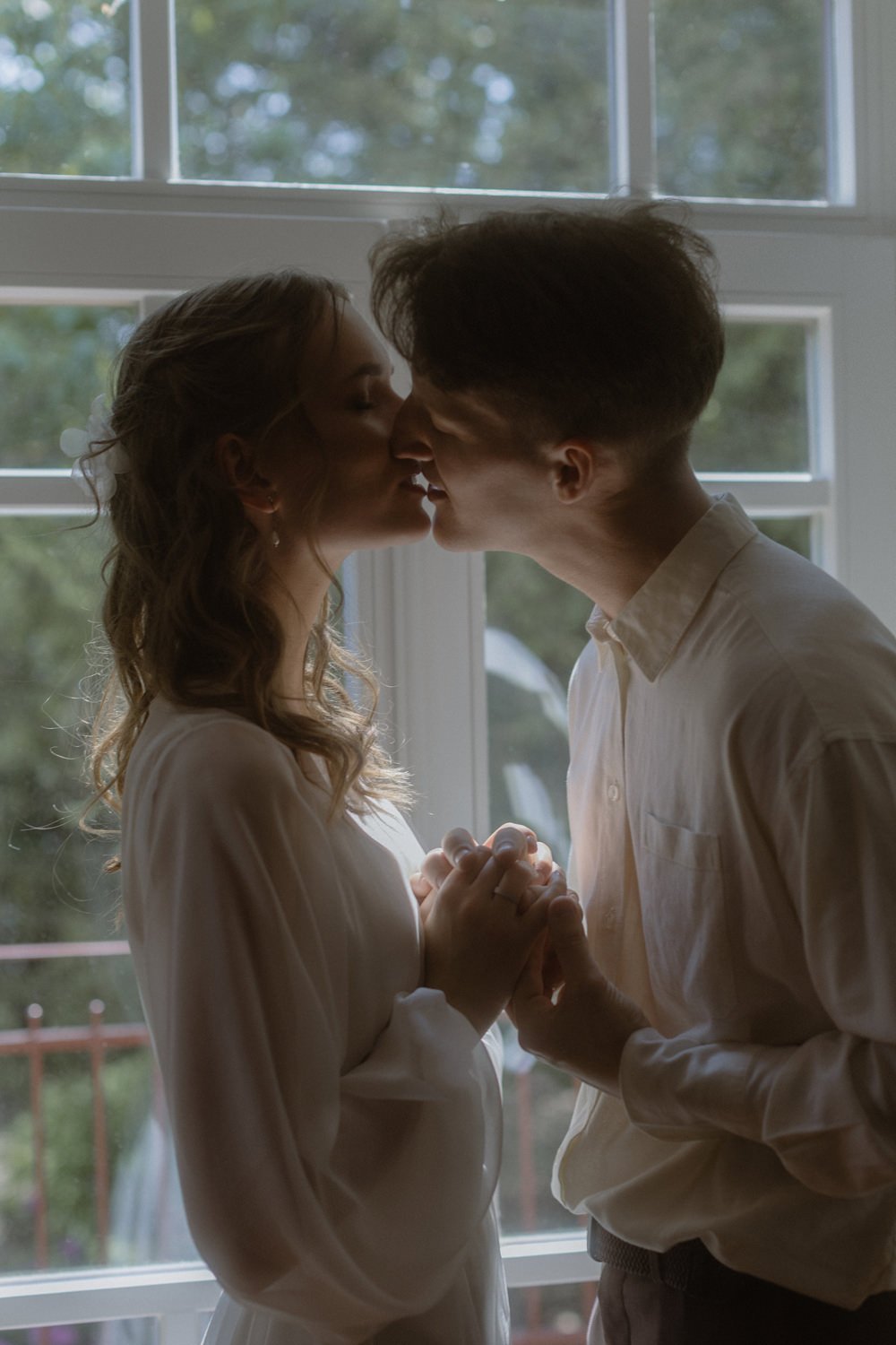 A man and woman in a sunlit room about to kiss, holding hands, with a window showing trees outside.