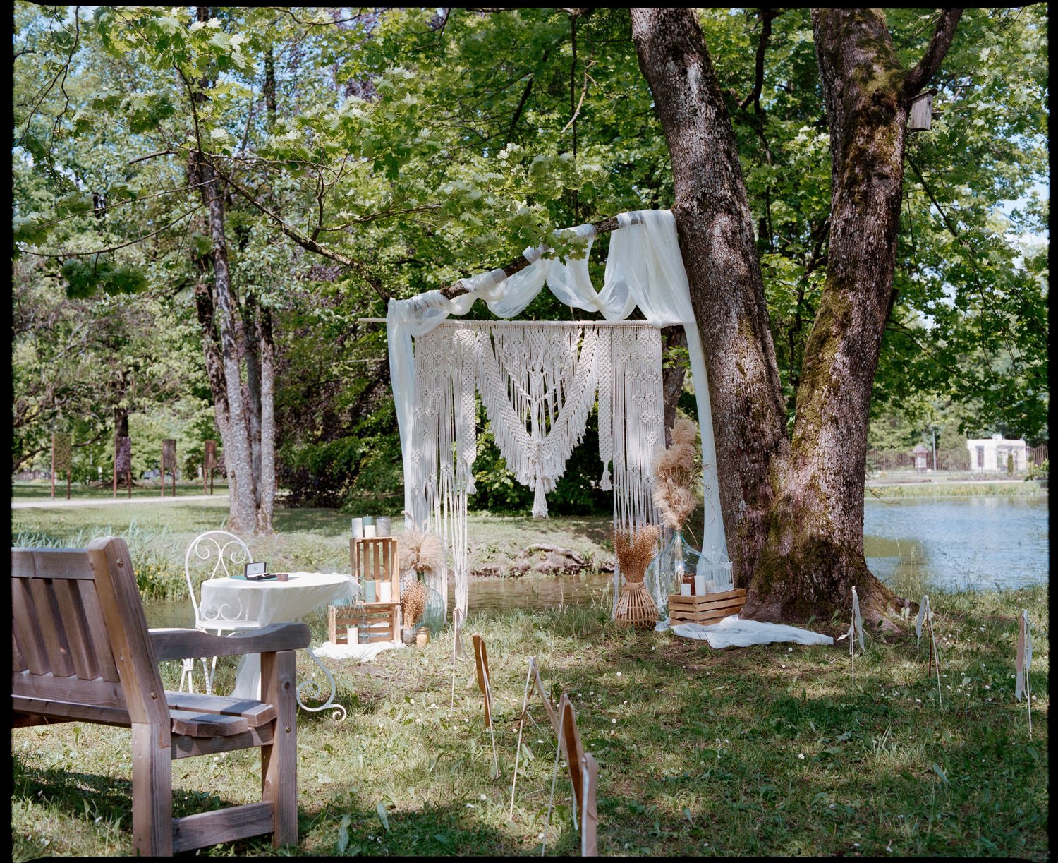 Bohemian outdoor wedding altar setup near a lake, with a macramé backdrop, white fabric draped over a tree branch, wooden and metal furniture, candles, decorative vases with dried flowers, and string lights, surrounded by greenery.
