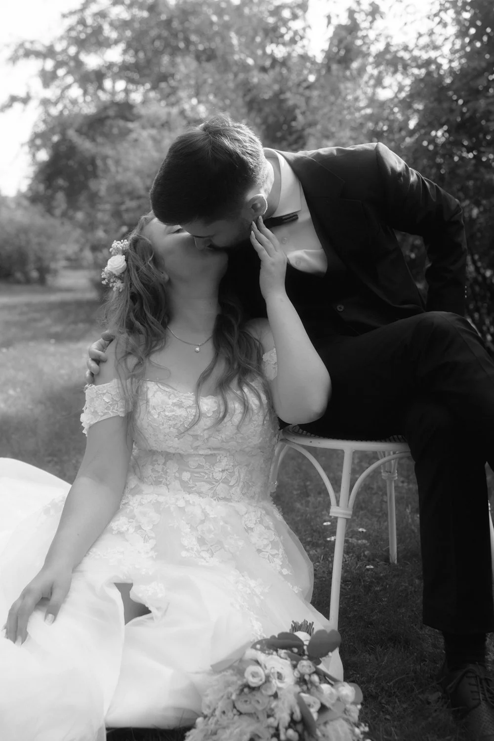 A black and white photo of a bride and groom sharing a kiss outdoors, with trees in the background. The bride is seated, wearing a wedding dress with lace details, and the groom is leaning over her, dressed in a tuxedo. The bride has flowers in her h