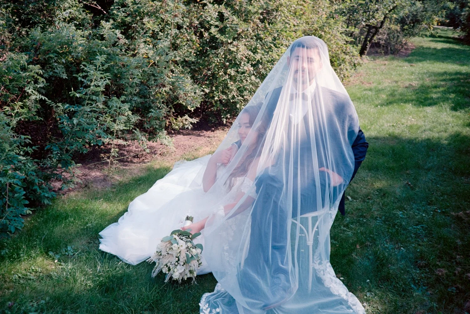A bride and groom seated outdoors on a sunny day, with a veil draped over both of them, covering their faces. The bride is holding a bouquet of flowers and resting against the groom, who has his arm around her. There are bushes and greenery in the ba