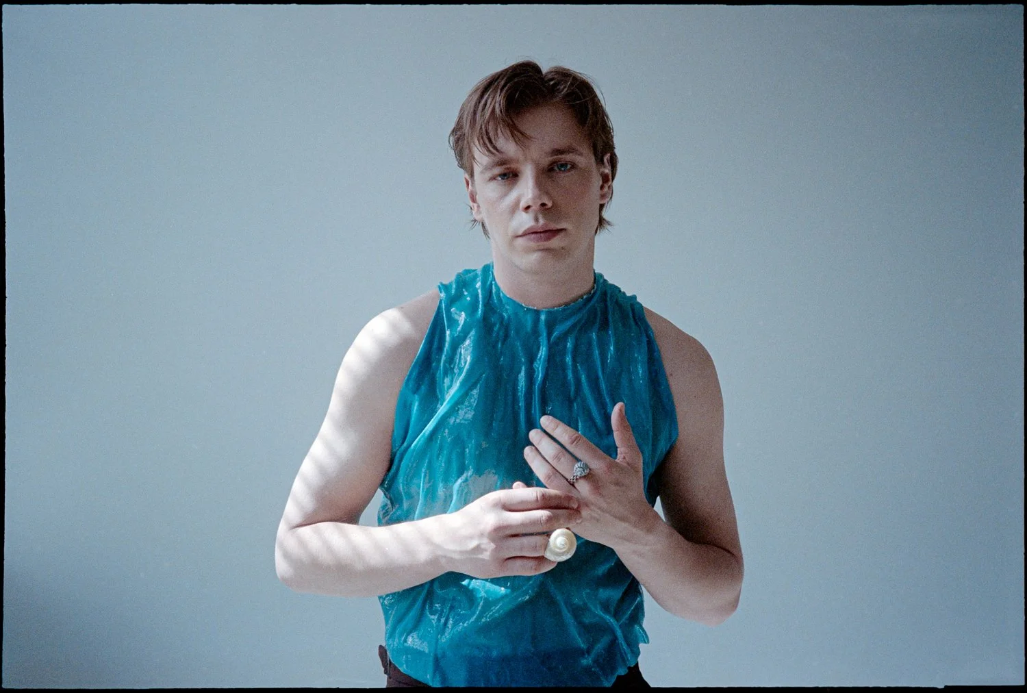 A young person with auburn hair and fair skin is standing against a plain gray wall. They are wearing a blue, shiny, sleeveless top and holding a pearl ring up over their chest with one hand while the other hand is placed over their heart. Their expr