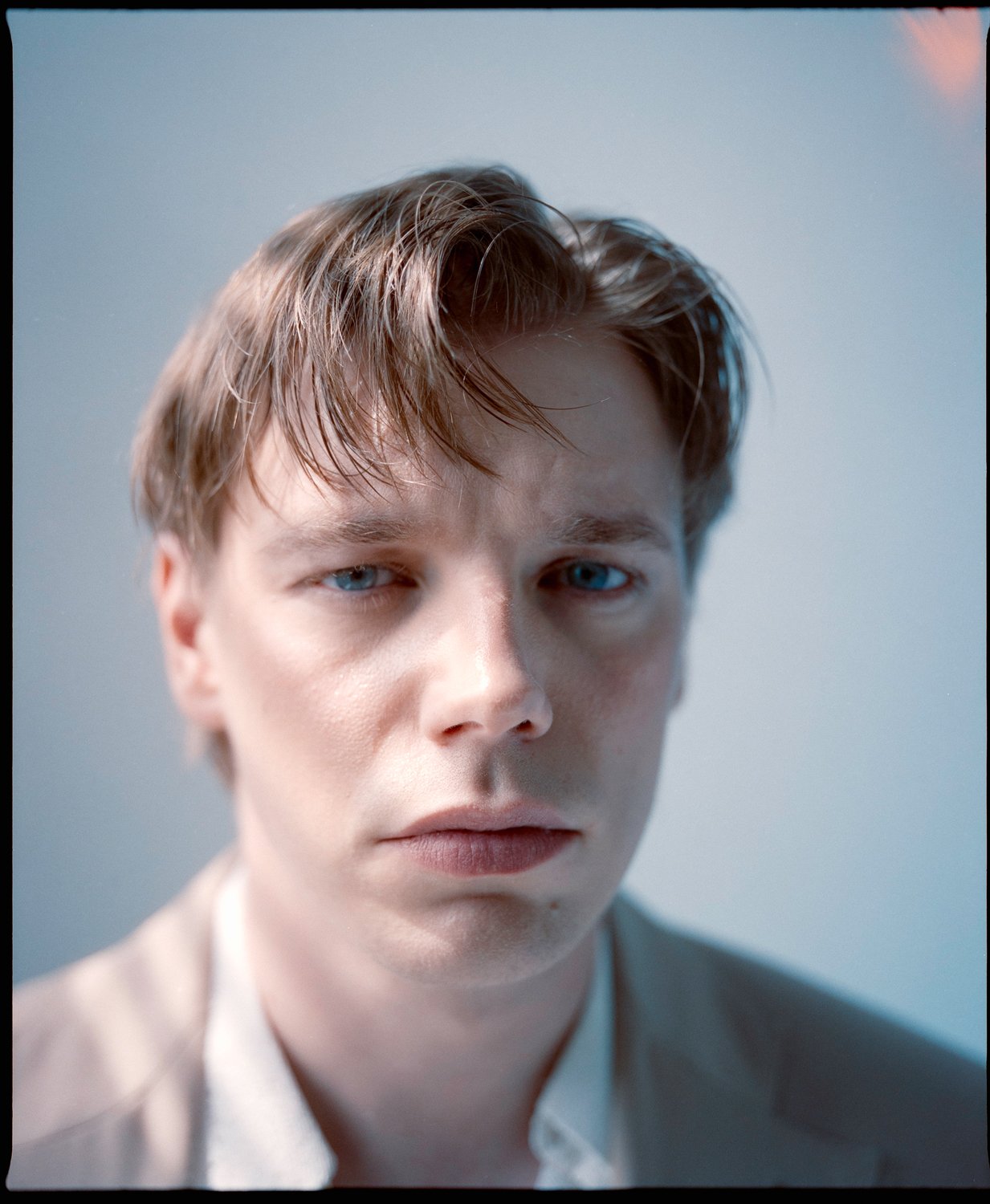 Close-up of a young man with light skin, blue eyes, and short brown hair, wearing a light-colored shirt, with a neutral expression and a plain background.