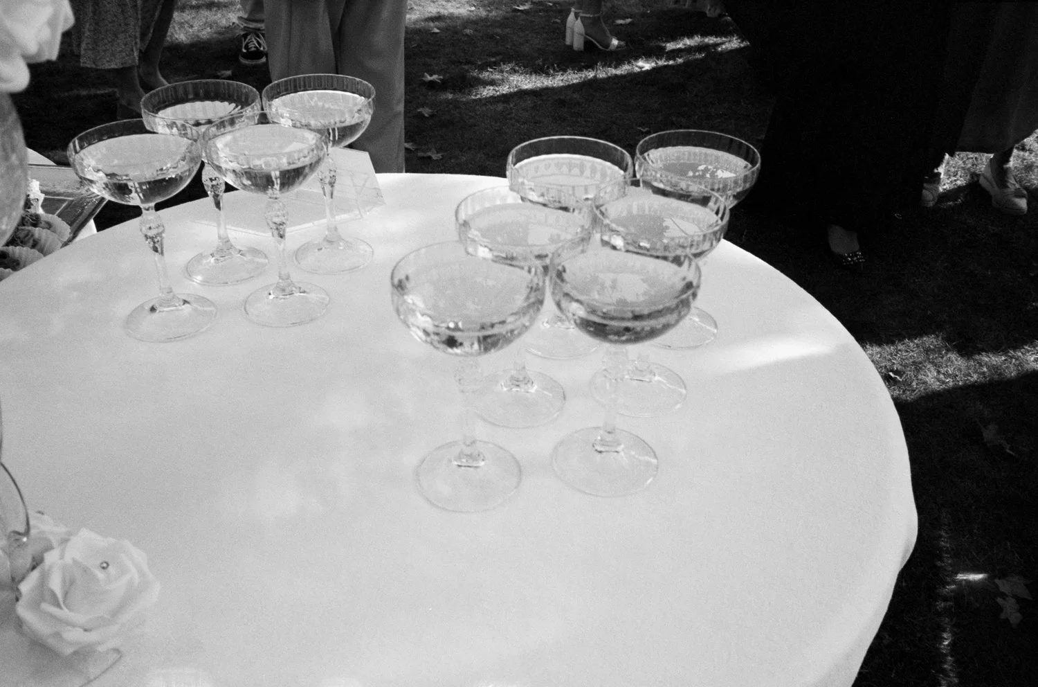 A table with 10 empty champagne glasses arranged on a white tablecloth, with people standing around in the background outside.