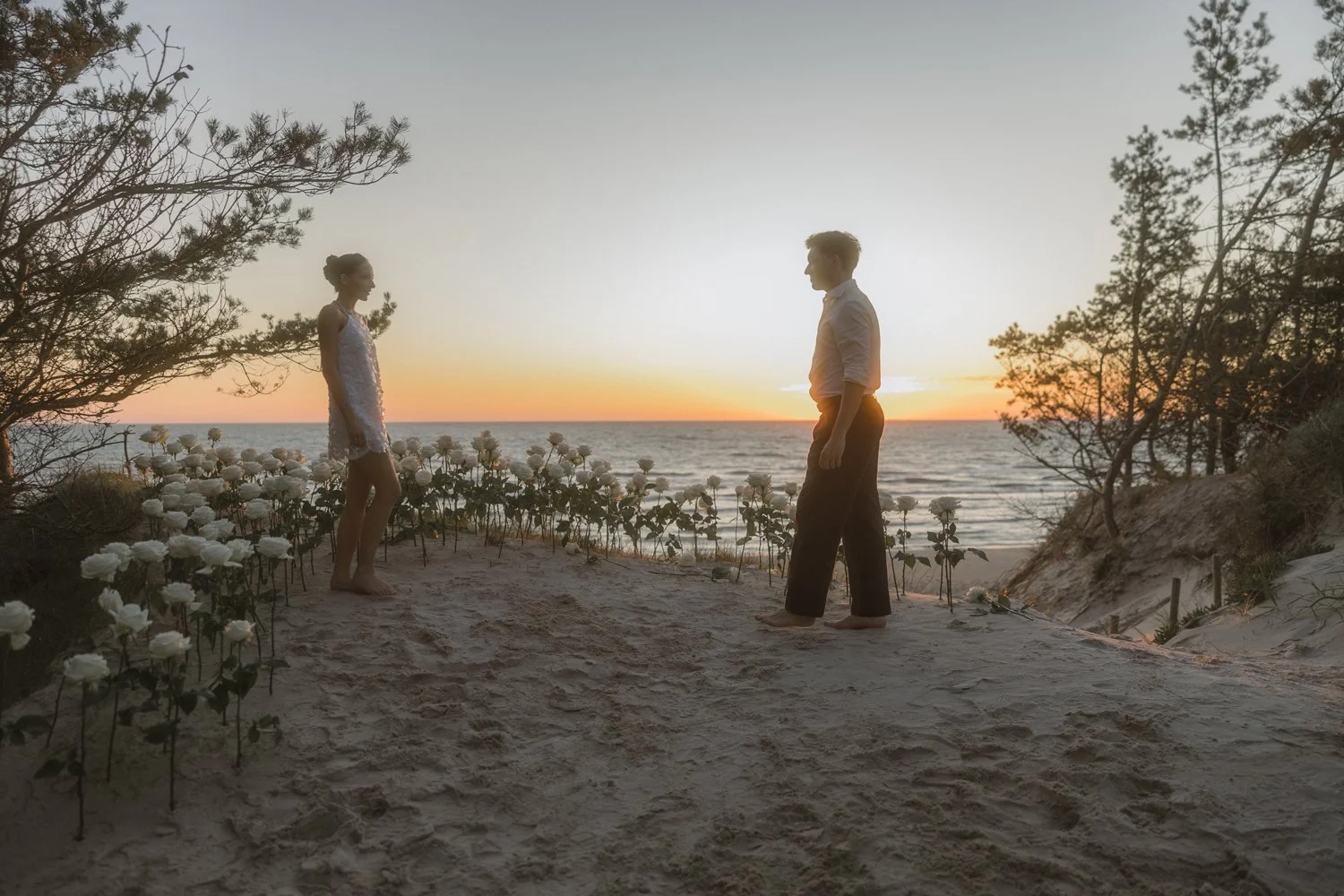 A man and woman stand facing each other on a sandy beach at sunset, surrounded by white flowers and trees, with the ocean in the background.