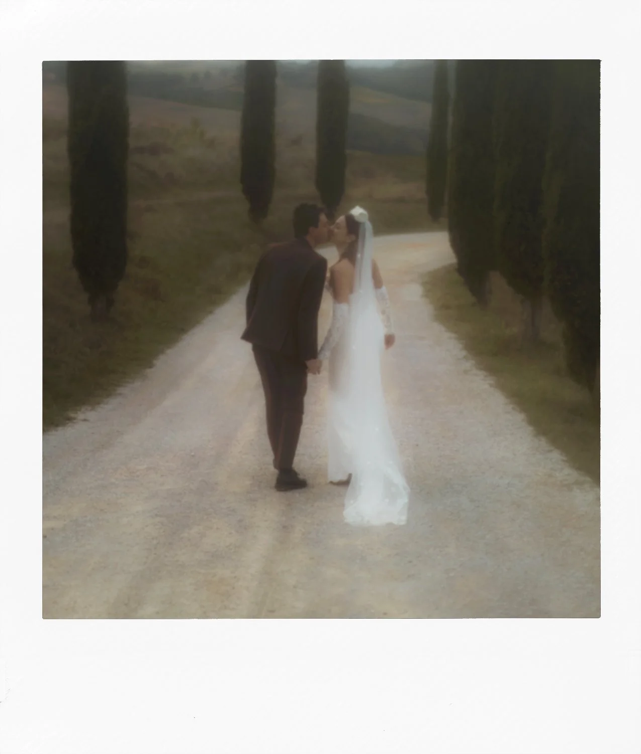 A bride and groom kissing on a dirt path lined with tall cypress trees.