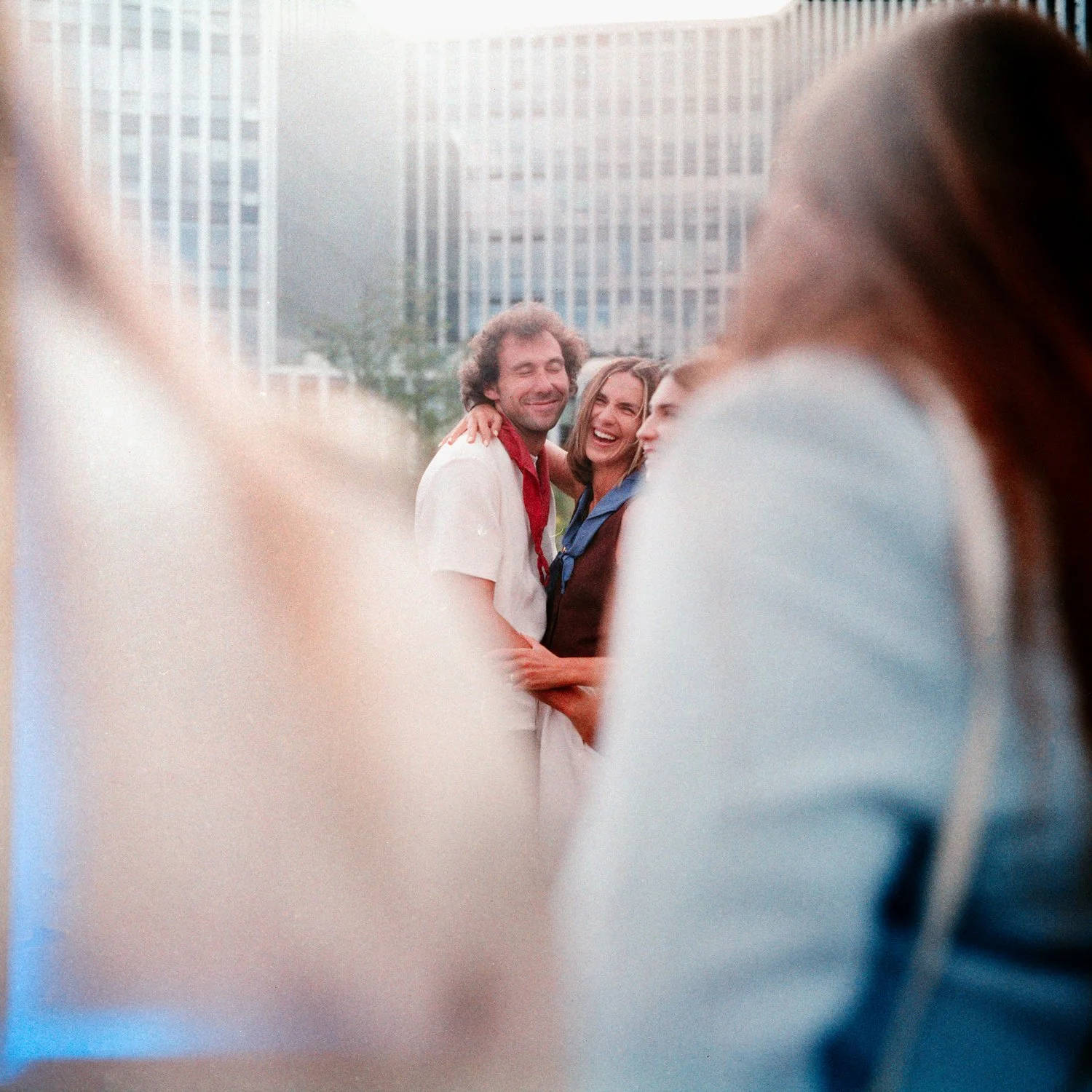 Two people smiling and hugging in an urban outdoor setting with tall buildings in the background, seen through a blurred foreground.