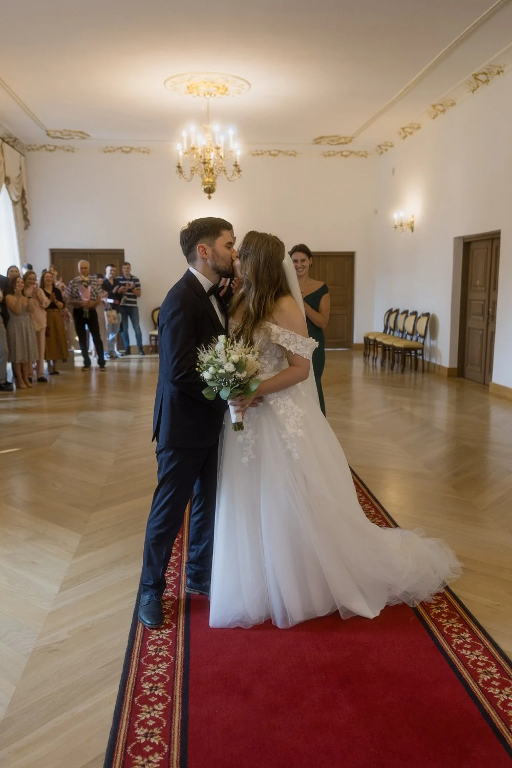 A bride and groom kissing during their wedding ceremony in an elegant hall with wooden floors and chandeliers, surrounded by wedding guests.
