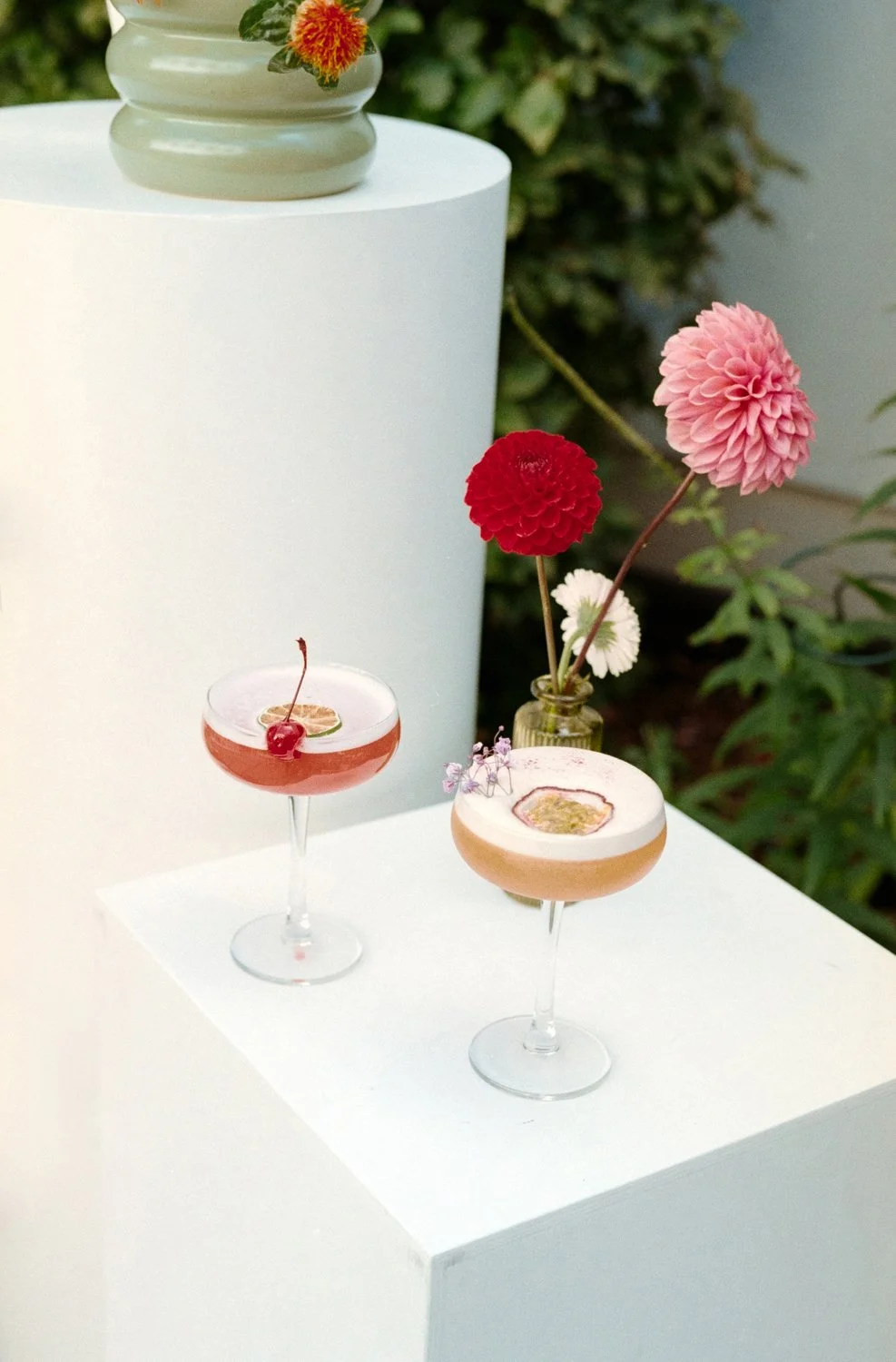 Two cocktails with flower garnishes on a white surface, with a flower arrangement in a small bottle in the background and blurred green foliage.
