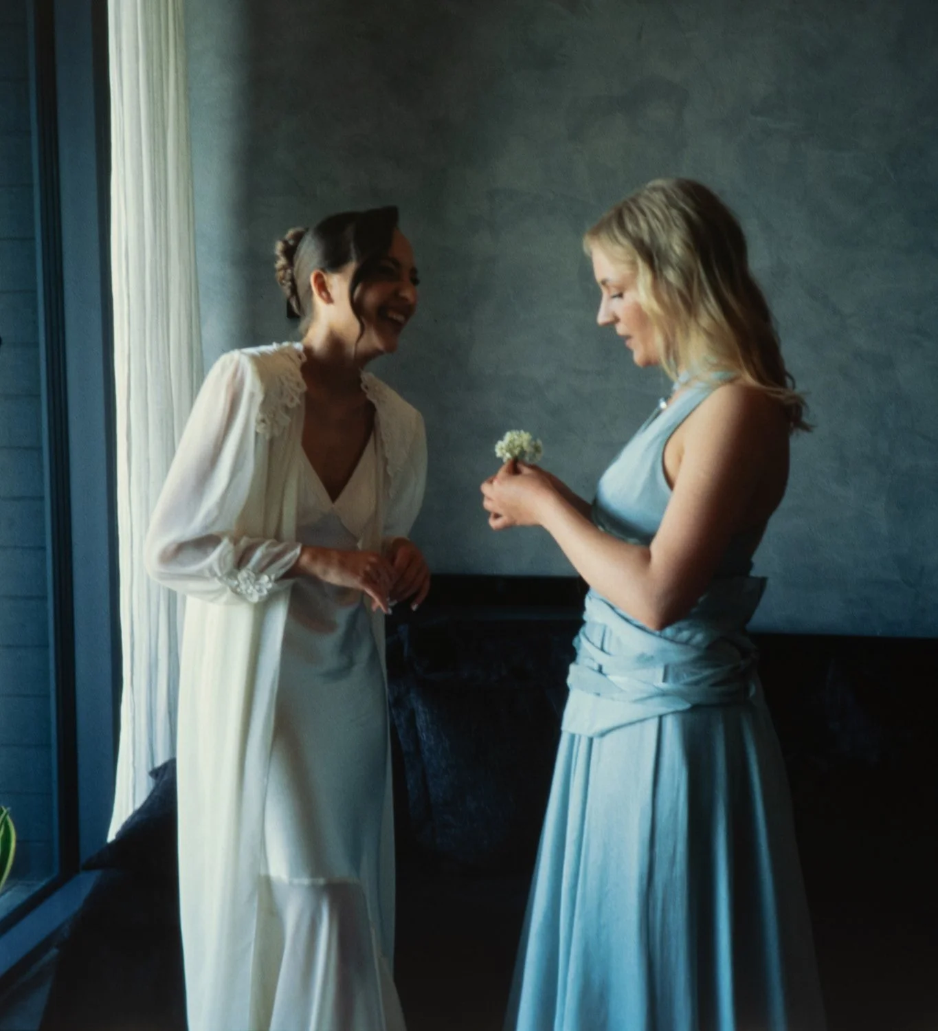 Two women, one in a white dress and the other in a light blue dress, smiling and interacting indoors with a gray textured wall in the background. The woman in blue is holding a small bouquet of white flowers.