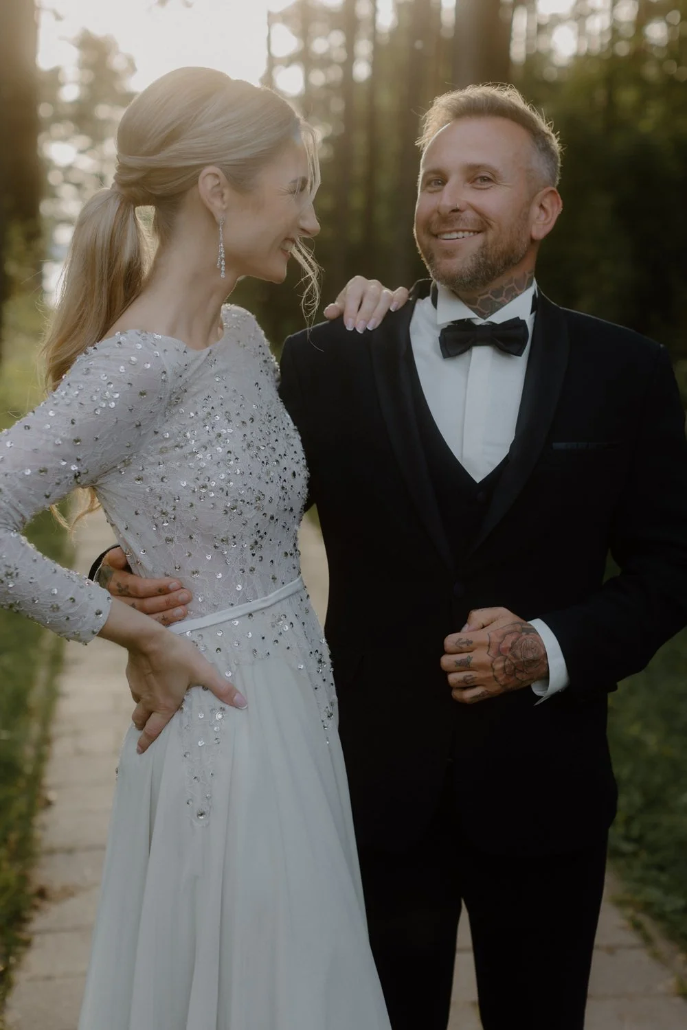 A bride and groom smiling and looking at each other outdoors during sunset, with trees in the background. The bride is wearing a long-sleeved, embellished wedding dress and the groom is dressed in a black tuxedo with a bow tie.