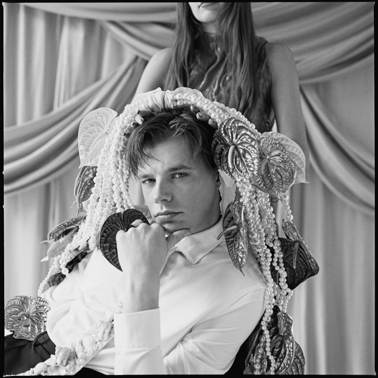 A young man with styled hair wearing a white shirt, sitting with his chin resting on his hand, surrounded by elaborate decorations including large leaves and pearl-like beads, with a woman standing behind, adjusting his headpiece, in front of a drape