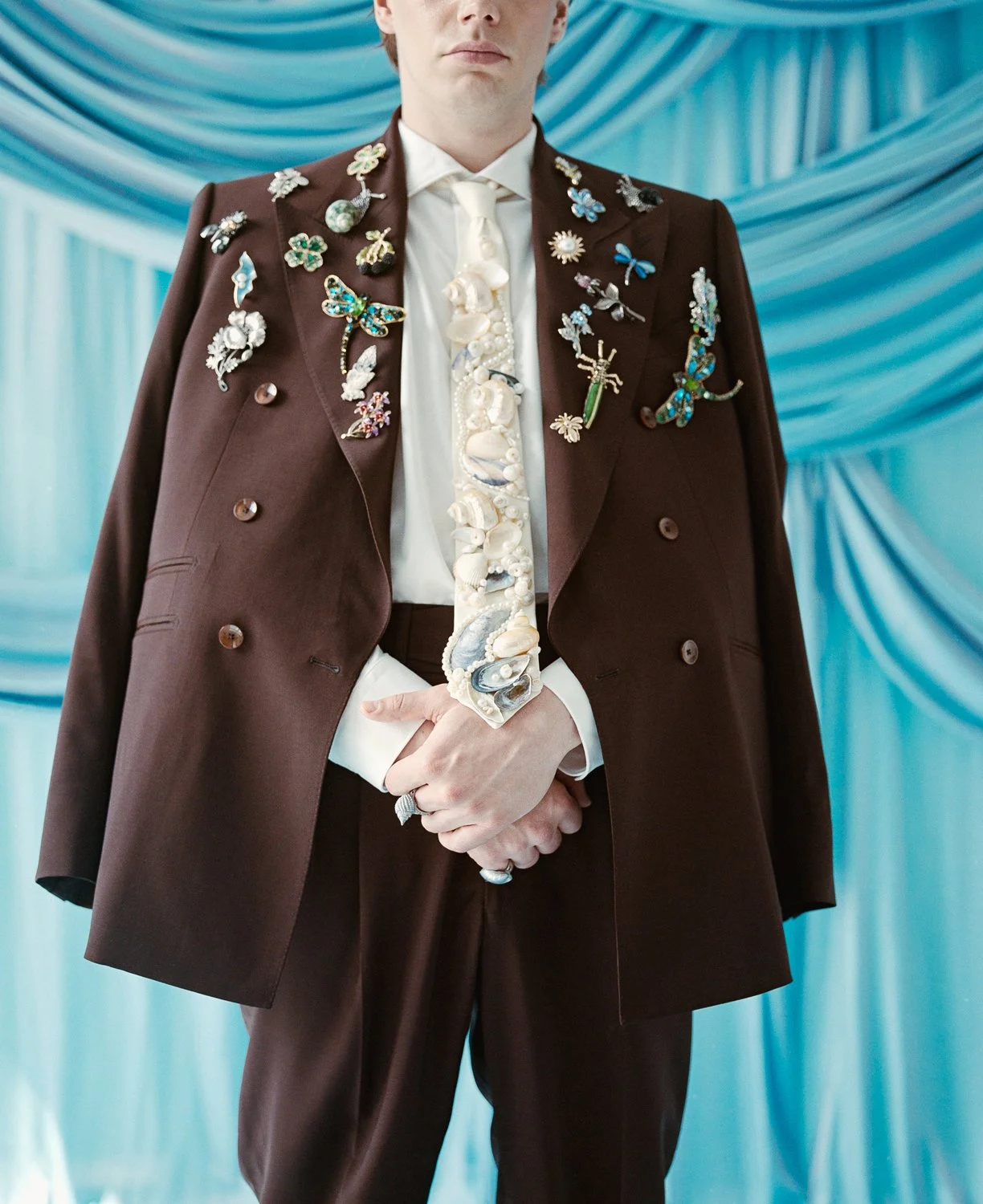 A person in a brown suit jacket adorned with various colorful butterfly and flower pins, wearing a white shirt, beige tie, and shells as a necklace, standing in front of blue drapery.