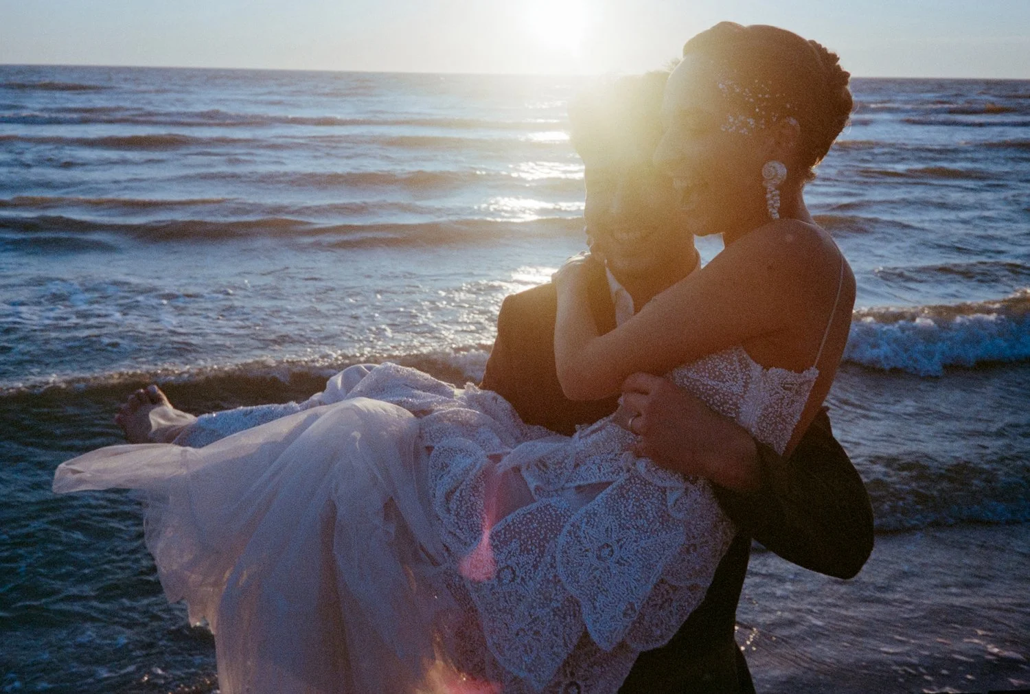 A woman in a dress holding a man in a tuxedo at the beach during sunset.