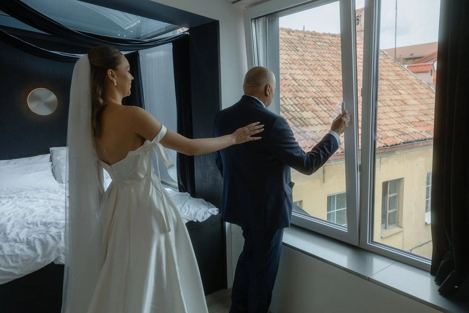 A bride in a white wedding dress with a veil and a groom in a suit looking out a window in a hotel room.