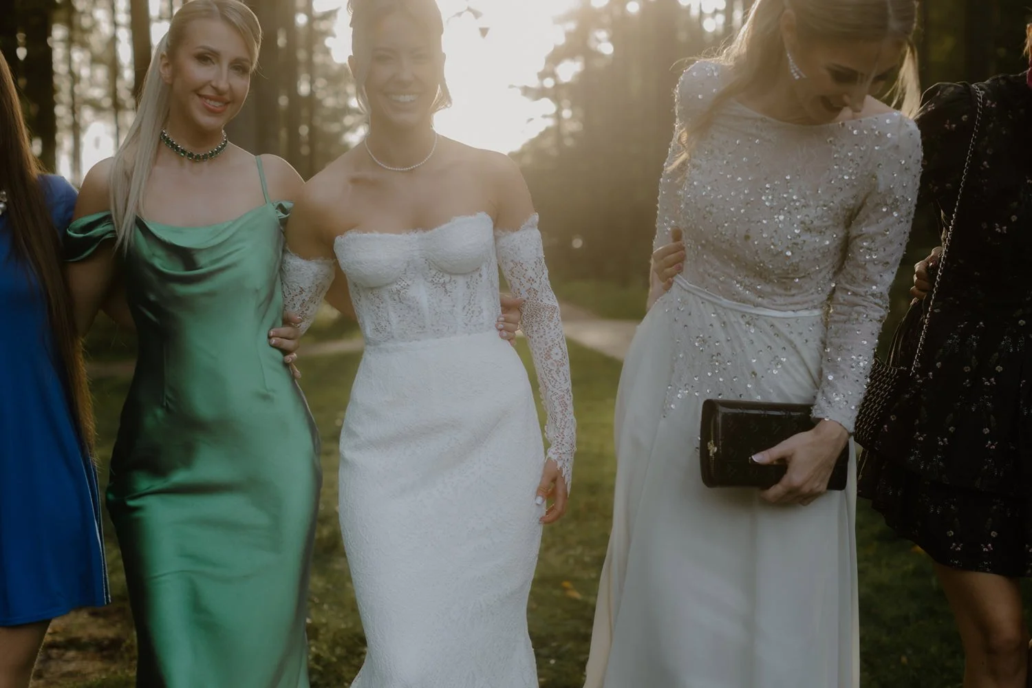 Group of women in formal dresses standing outdoors in a forest, smiling and posing for a photo.